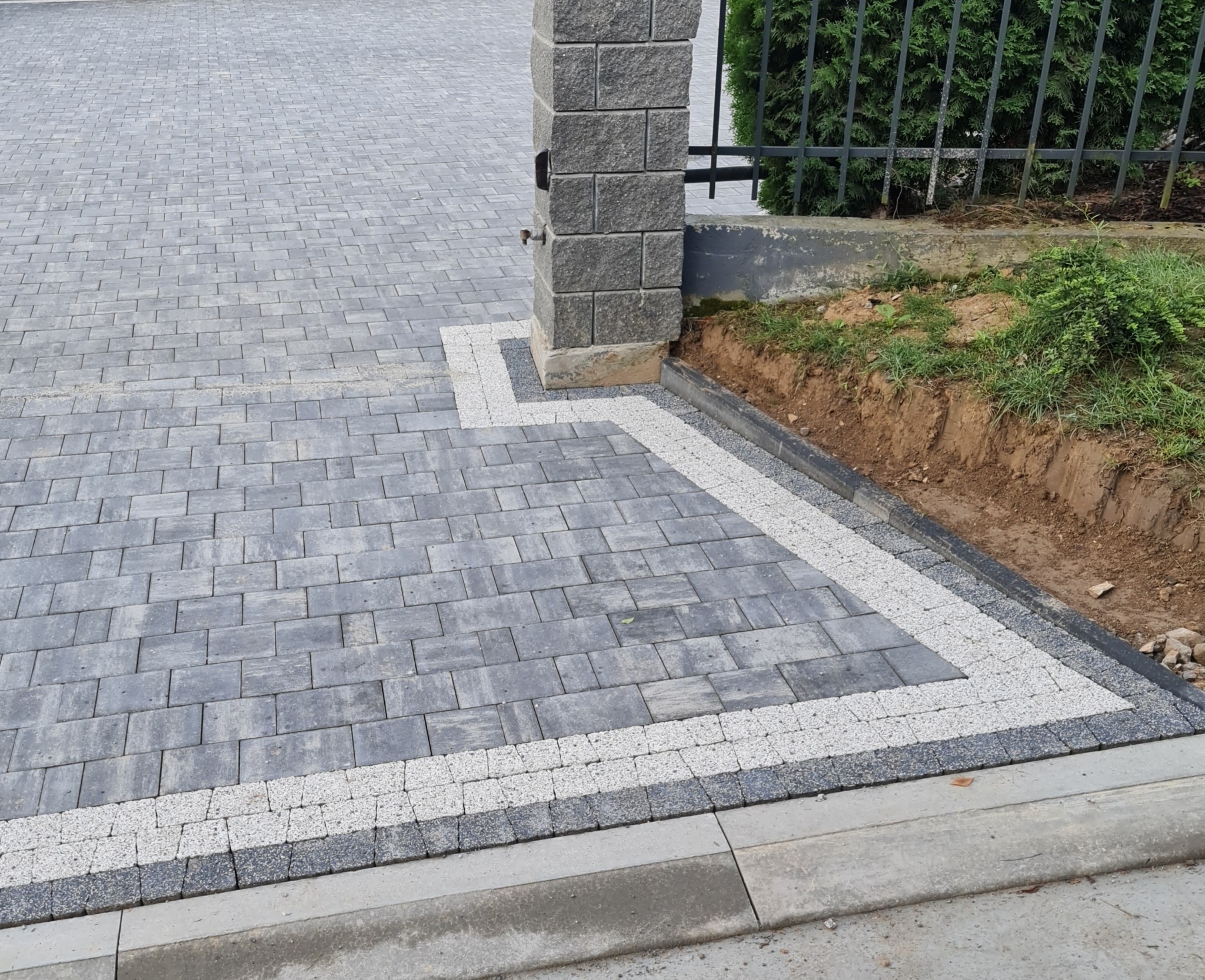 Szara kostka brukowa ułożona wzorem z obramowaniem z jasnych kamieni, przylegająca do kamiennego słupka ogrodzeniowego i krawężnika.