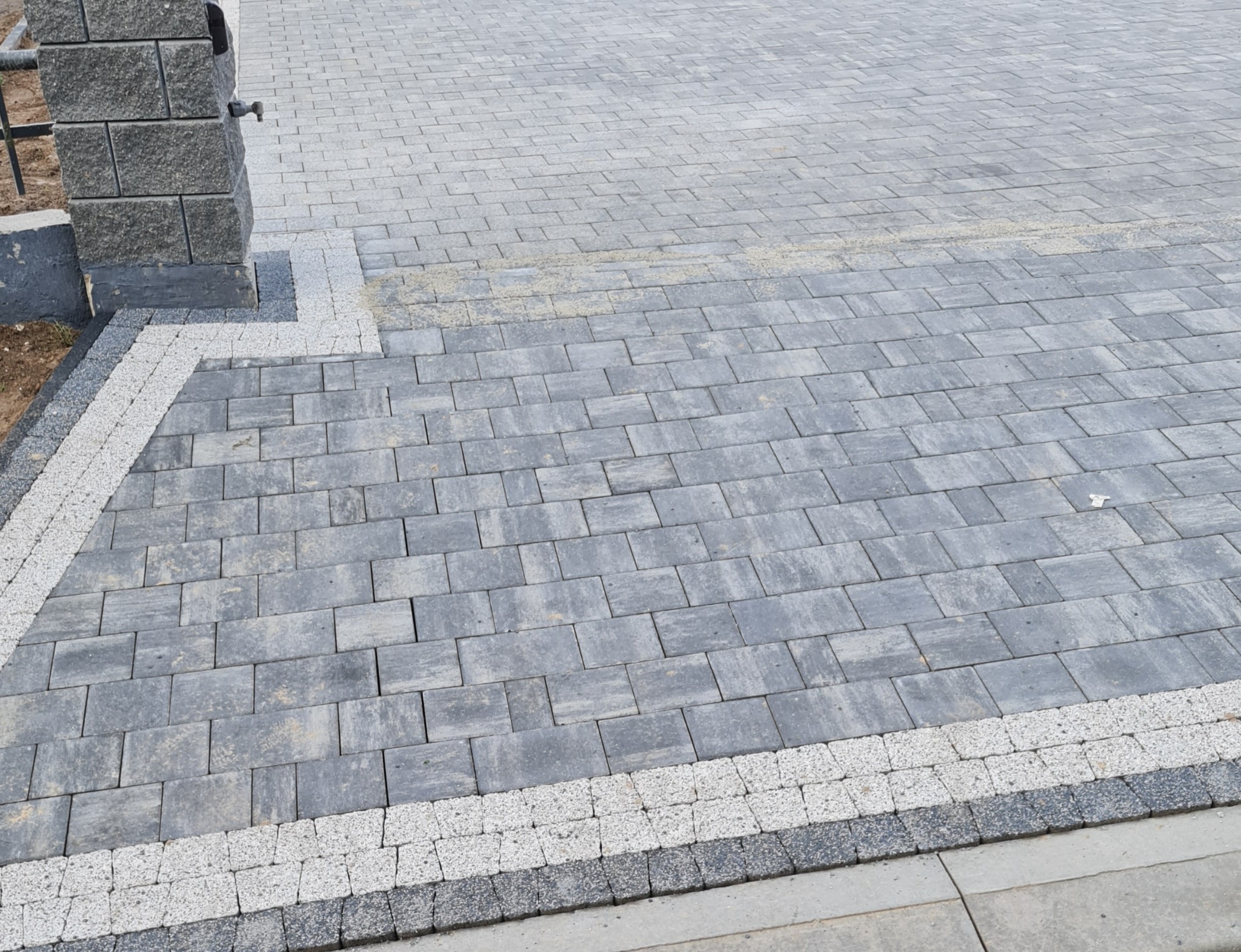 Szara kostka brukowa ułożona na podjeździe, obrzeża z jasnej kostki granitowej, widoczny słupek ogrodzeniowy z szarej kostki.