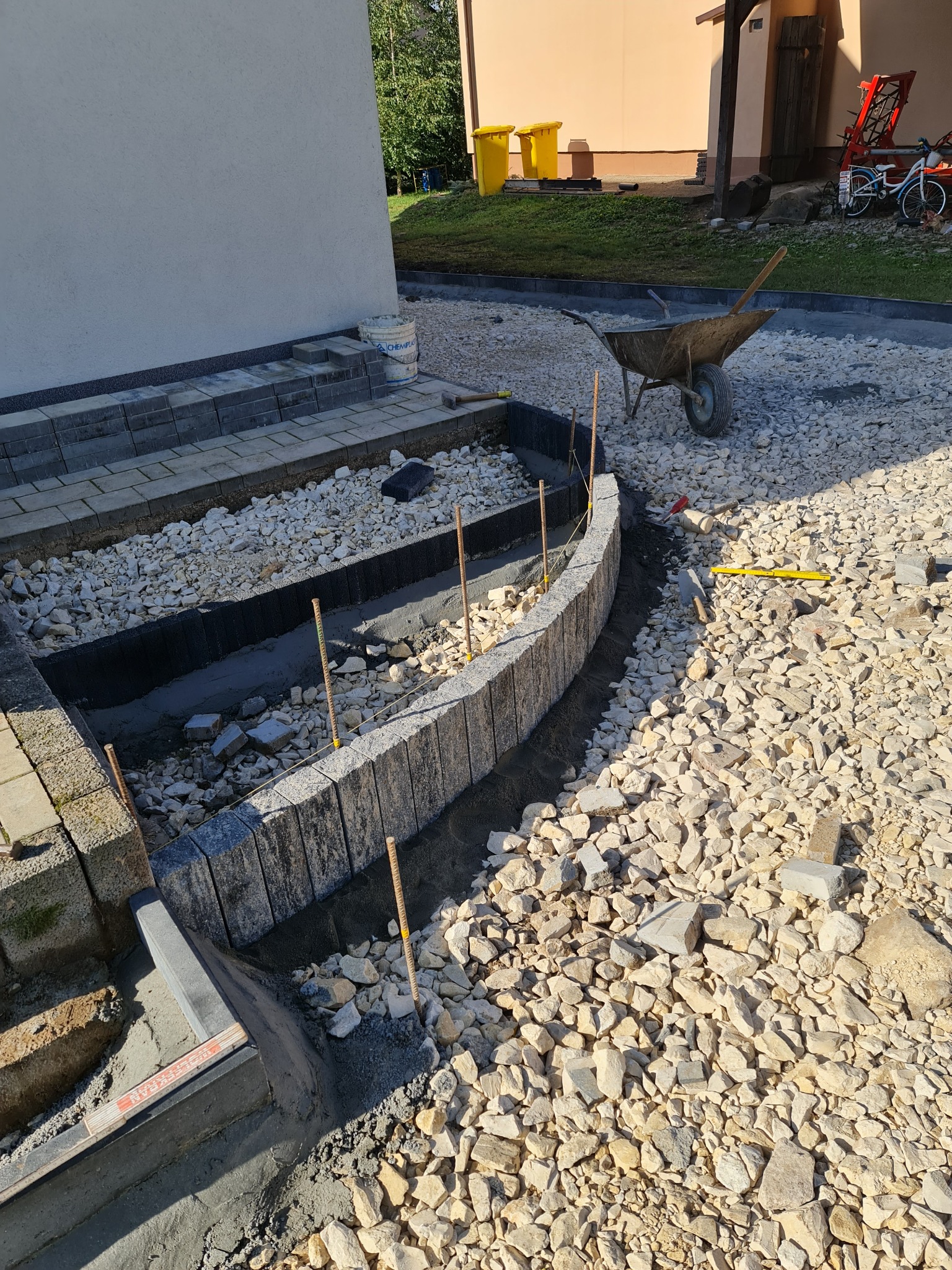 Budowa obrzeża rabaty z kostki granitowej w Proszowicach. Widoczne szare kostki ułożone w łuk, wypełnienie z białych kamieni, narzędzia budowlane i taczka w tle.