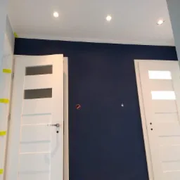 Wnętrze w trakcie remontu: świeżo pomalowana granatowa ściana, białe drzwi z szybami zabezpieczone żółtą taśmą malarską, widoczne otwory po gniazdkach elektrycznych.