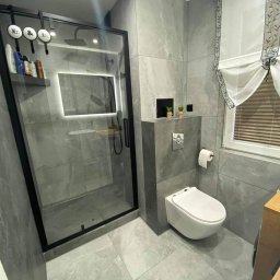 MarqoBUDS - Nowoczesna łazienka z szarymi płytkami imitującymi marmur, kabiną prysznicową typu walk-in z czarną ramą i podświetlanym lustrem oraz wiszącą toaletą.