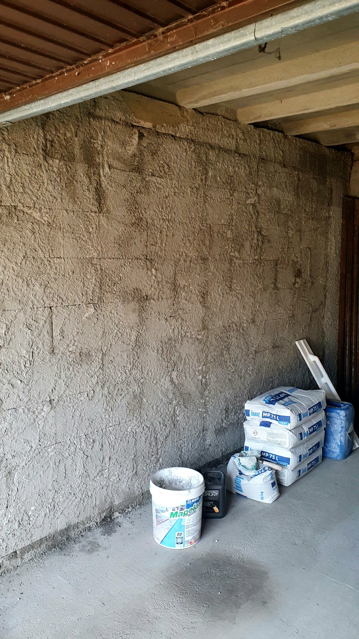 Ściana wewnętrzna po nałożeniu tynku strukturalnego, obok stoją worki z zaprawą Knauf MP 75 L, wiadro z gruntem Mapelas Royal Grunt i czarny kanister.