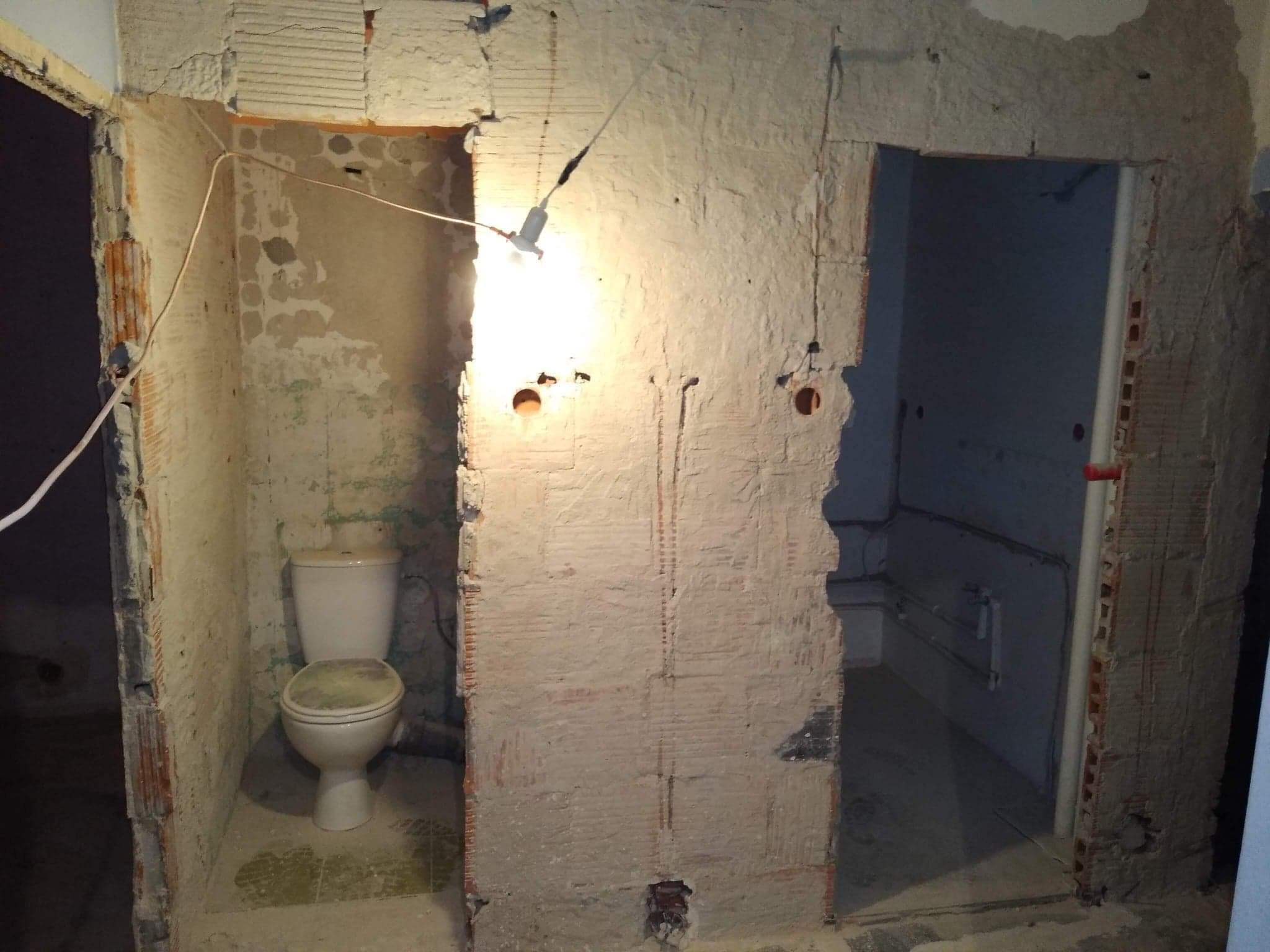 Łazienka w trakcie demontażu starej glazury, widoczne ślady po kuciu płytek na ścianach, surowa instalacja elektryczna z żarówką, muszla klozetowa w narożniku pomieszczenia.