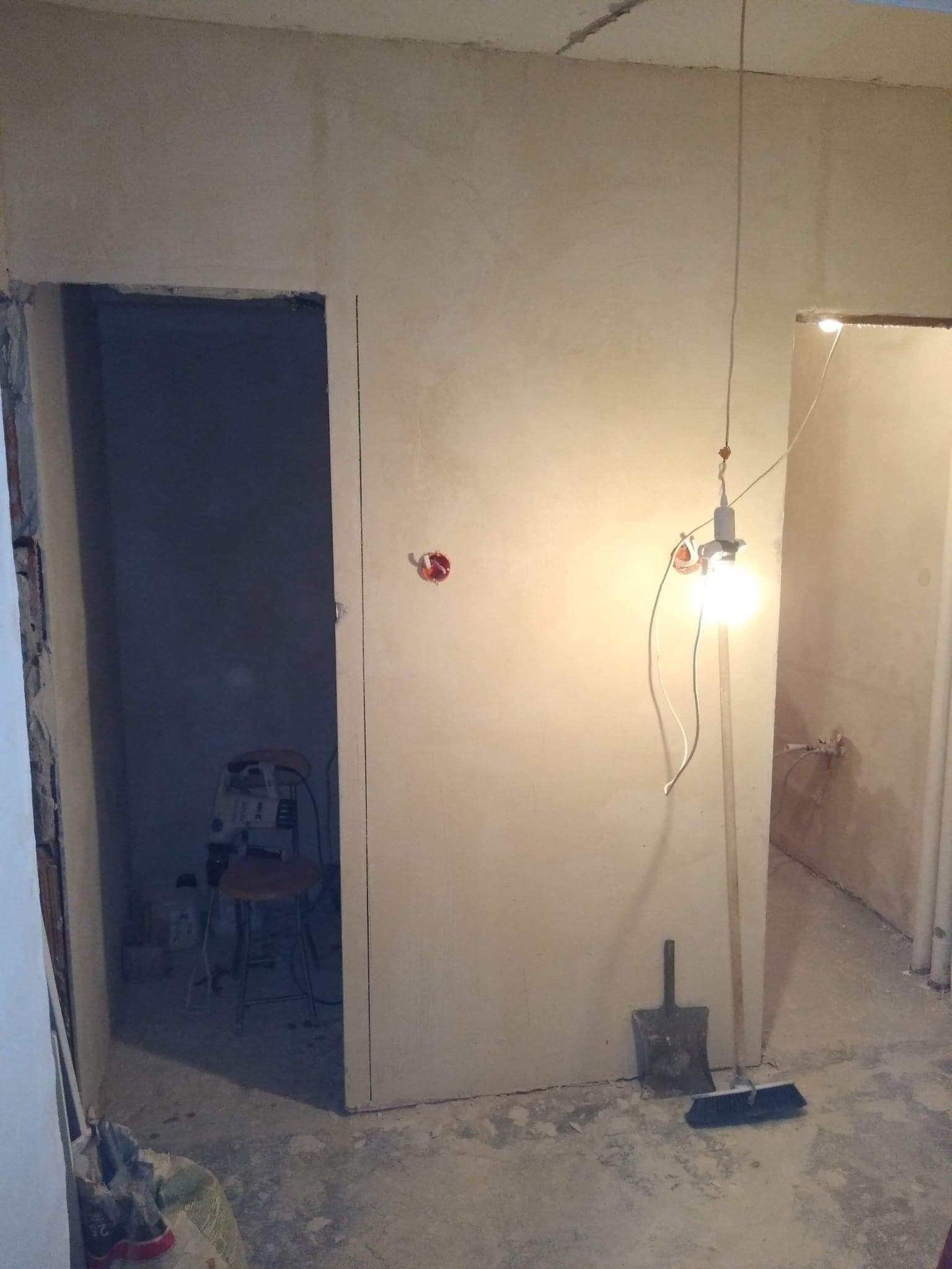 Surowe wnętrze mieszkania w trakcie remontu: widoczne ściany po tynkowaniu, prowizoryczne oświetlenie żarówką, otwory drzwiowe, narzędzia i materiały budowlane na podłodze.