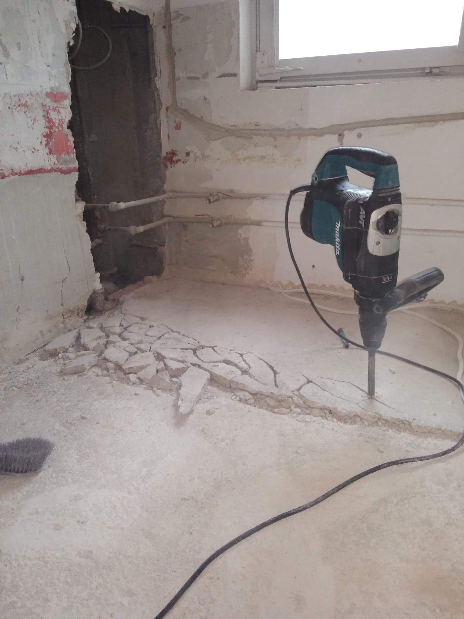 Kucie posadzki w trakcie prac remontowych, widoczny młot udarowy Makita, fragmenty gruzu na podłodze oraz odsłonięte rury w ścianie.