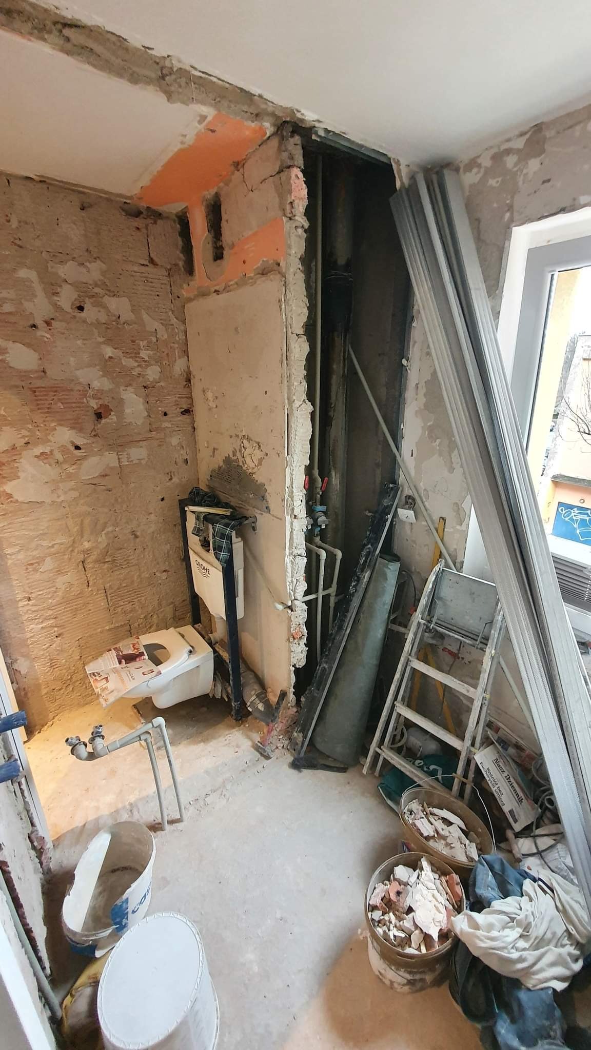 Demontaż w łazience: odsłonięte rury, stelaż podtynkowy WC Grohe, gruz w wiadrach, metalowe profile i drabina w trakcie prac remontowych.