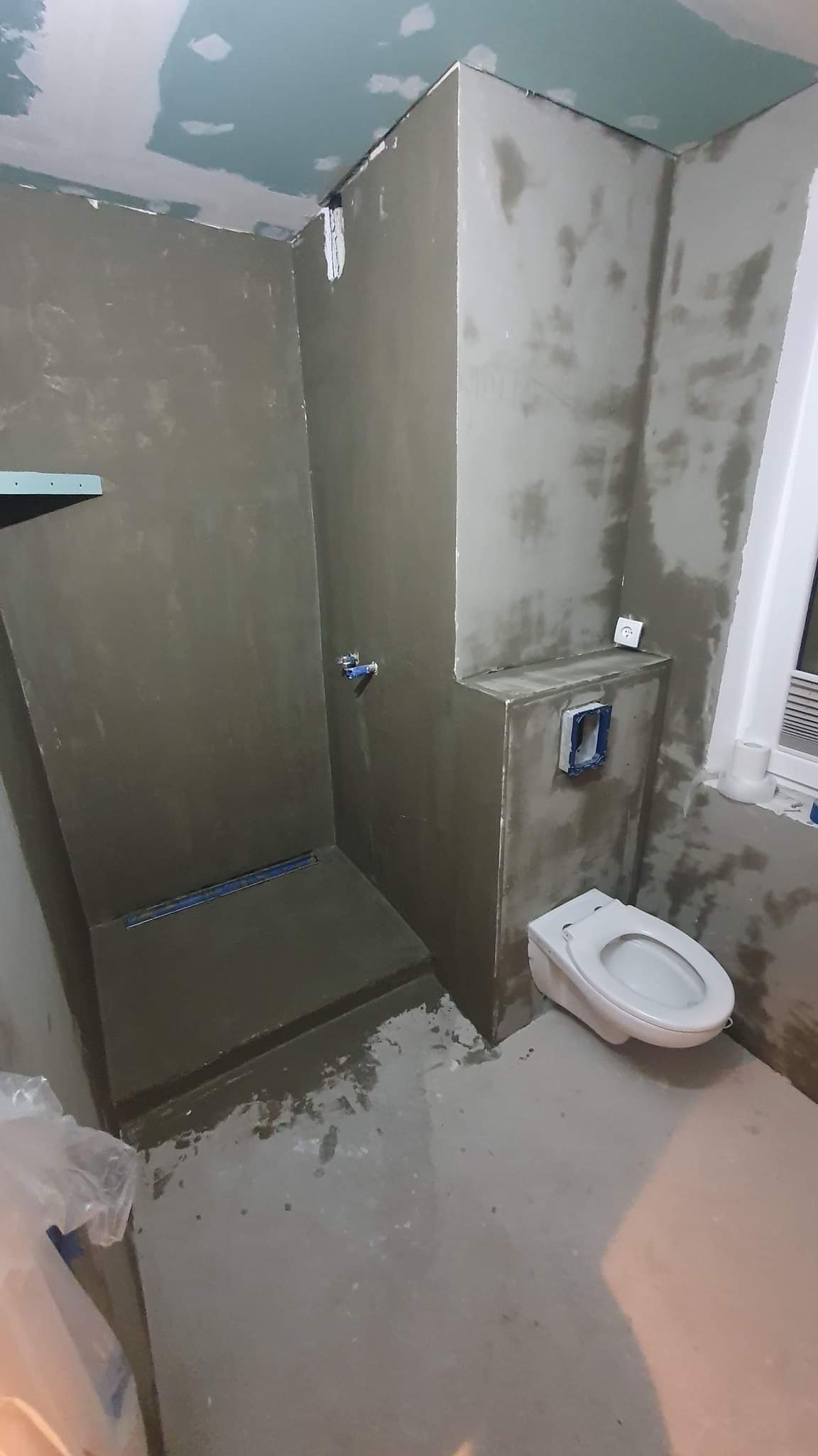 Surowy stan łazienki w trakcie prac: ściany pokryte szarym tynkiem, widoczny odpływ liniowy w brodziku, podwieszana toaleta, oraz gniazdko elektryczne na ścianie.