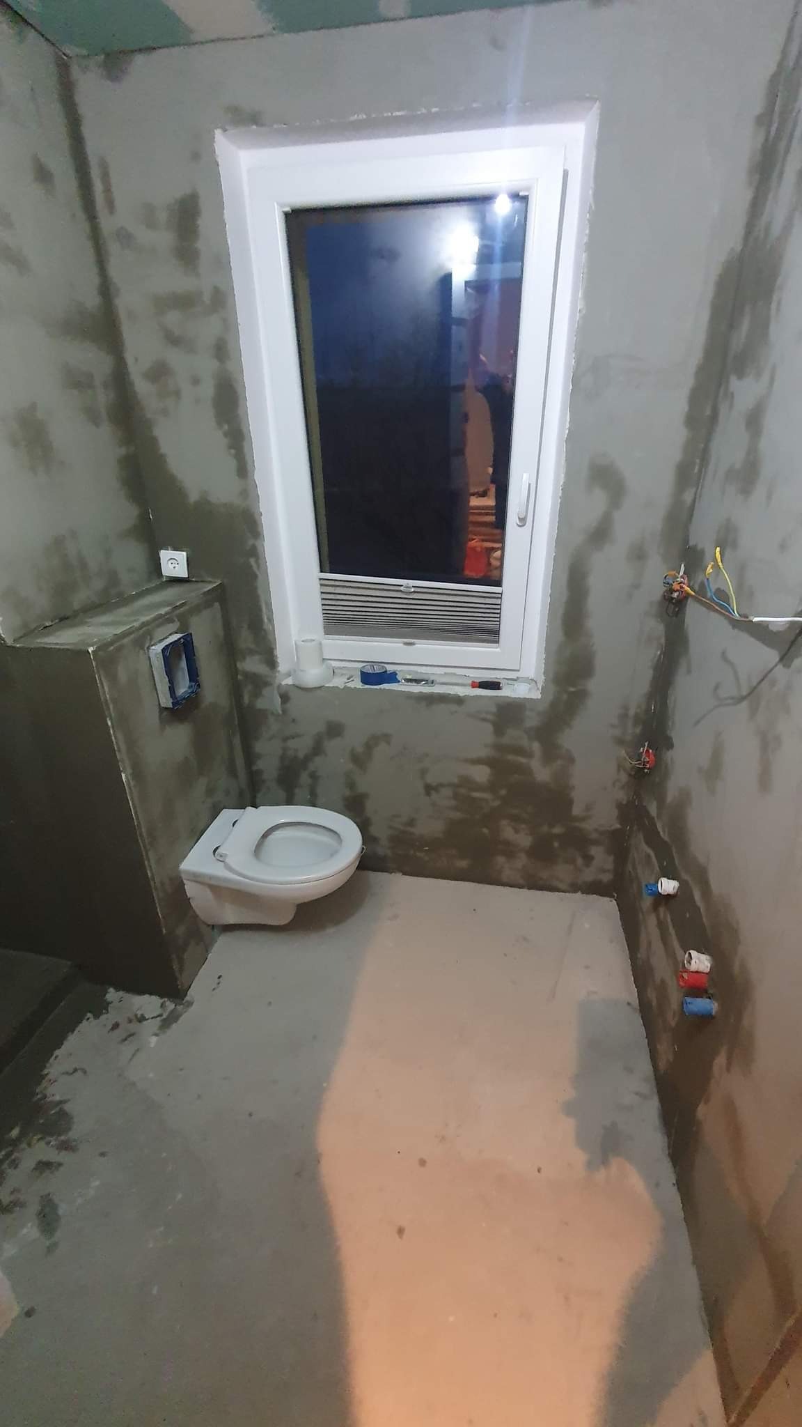 Surowa łazienka w trakcie prac: zamontowana podwieszana toaleta, ściany w szarym tynku, wystające przewody elektryczne i rury hydrauliczne, okno z roletą w tle.