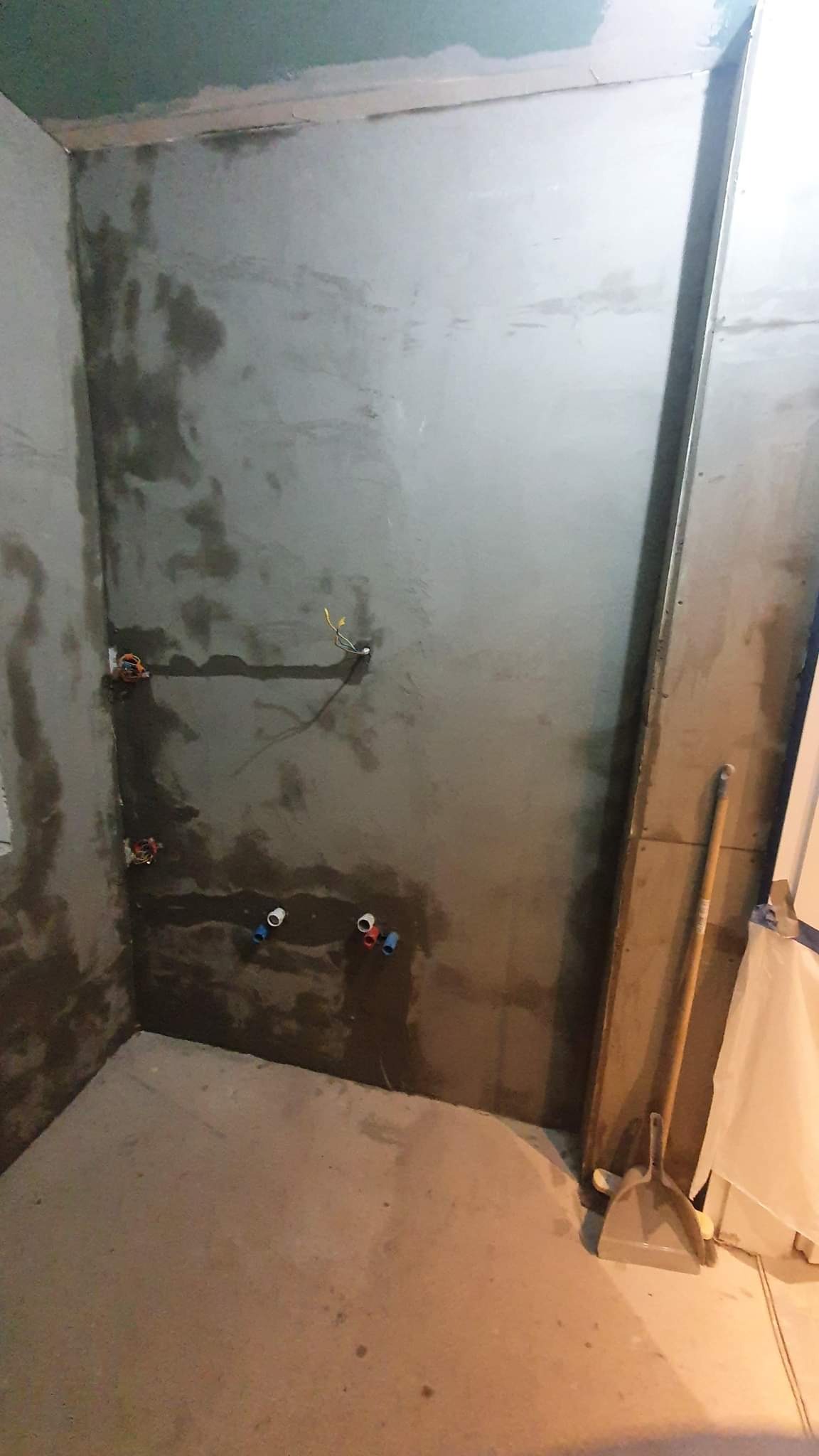 Surowa ściana w trakcie przygotowania do wykończenia w łazience, widoczne instalacje wodne i elektryczne, obok ściany oparta zmiotka i szufelka.