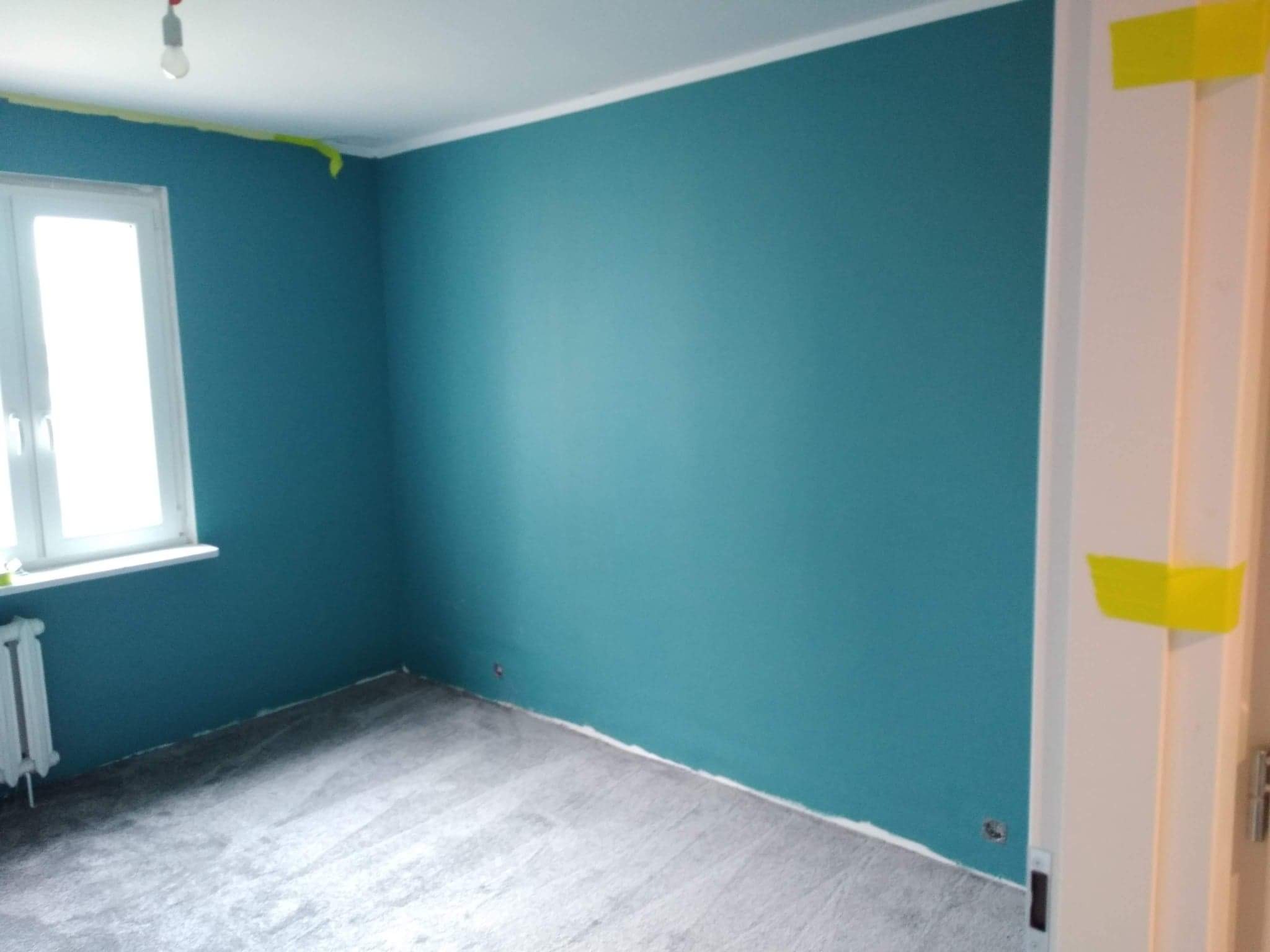 Pomieszczenie po malowaniu ścian na turkusowo, z widocznym zabezpieczeniem taśmą malarską przy ościeżnicy i suficie, szara podłoga.