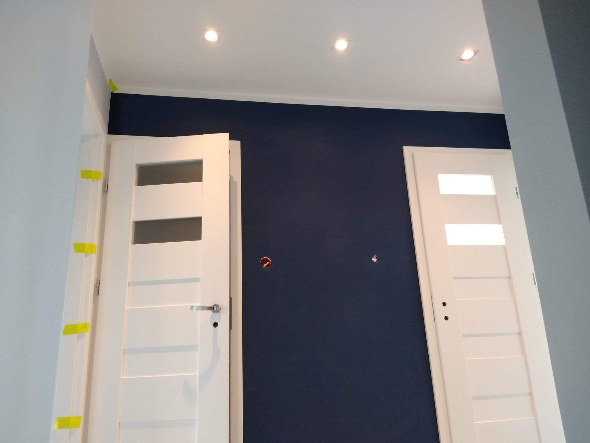 Wnętrze w trakcie remontu: świeżo pomalowana granatowa ściana, białe drzwi z szybami zabezpieczone żółtą taśmą malarską, widoczne otwory po gniazdkach elektrycznych.