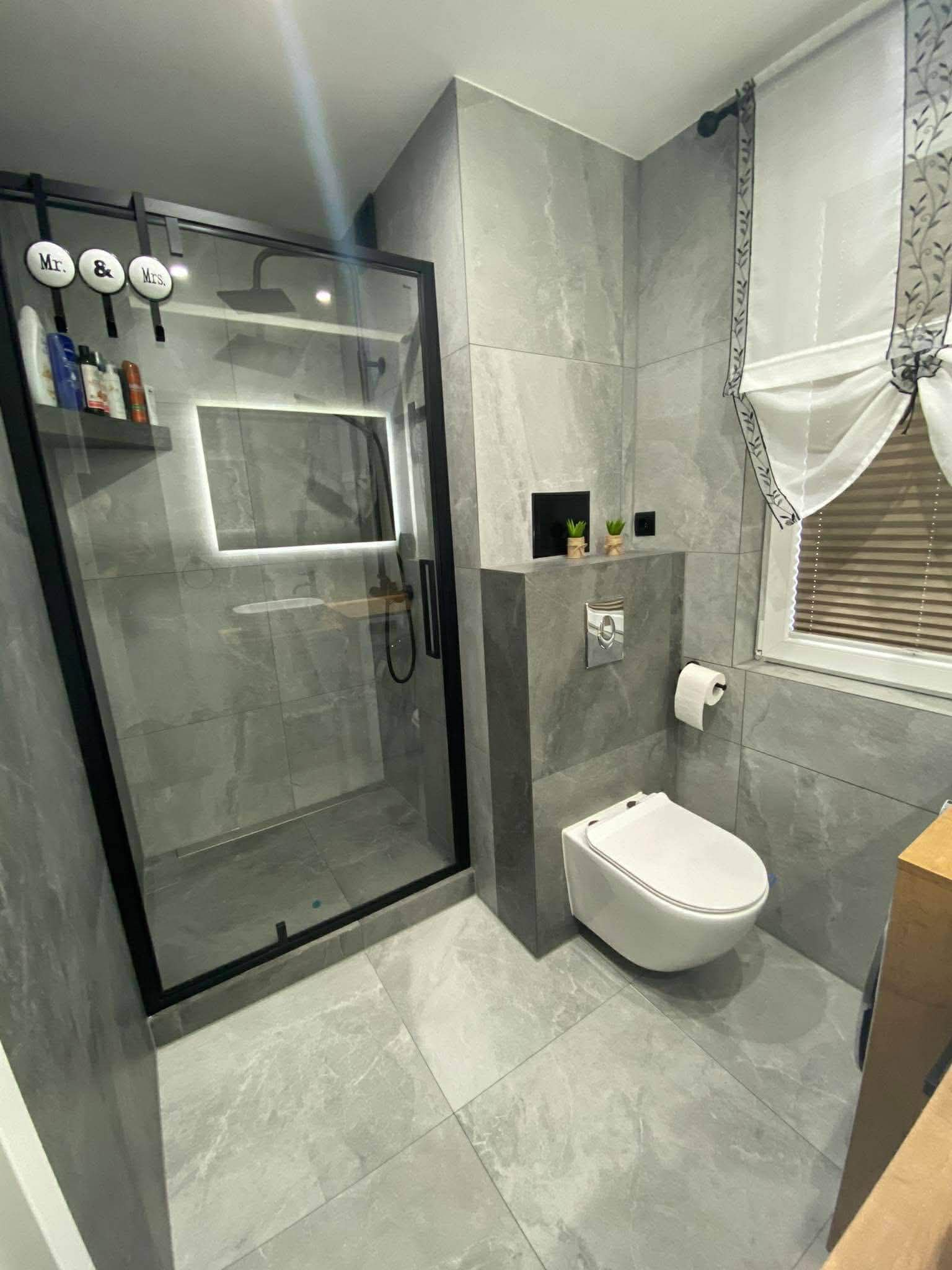 Nowoczesna łazienka z szarymi płytkami imitującymi marmur, kabiną prysznicową typu walk-in z czarną ramą i podświetlanym lustrem oraz wiszącą toaletą.