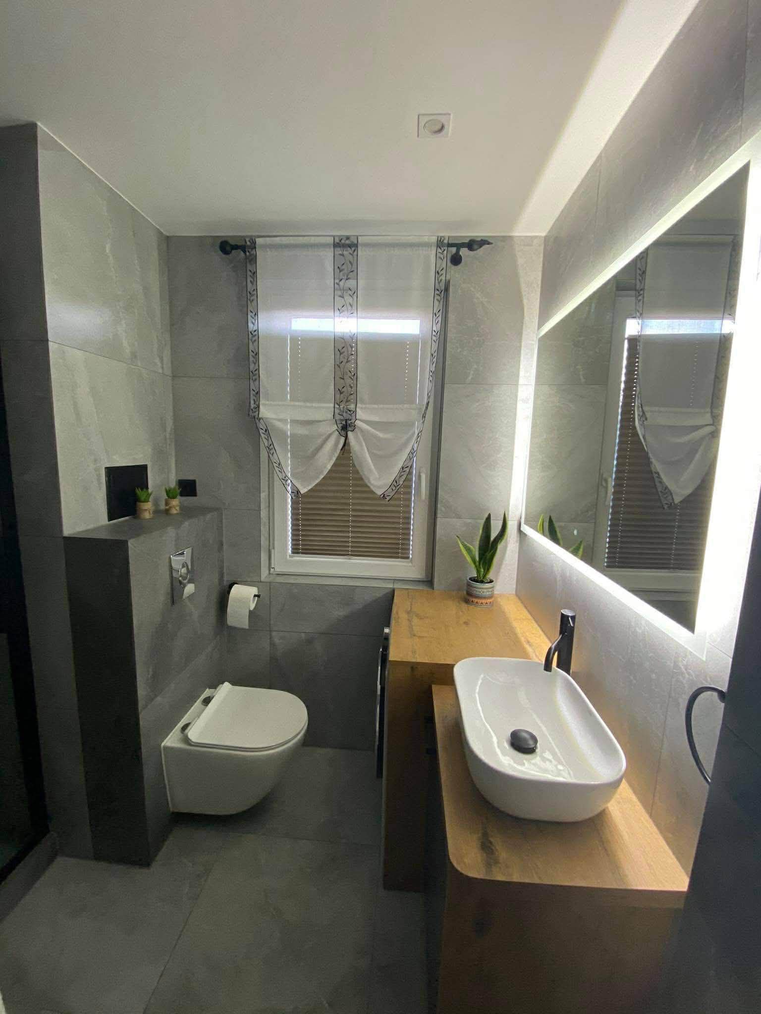Nowoczesna łazienka z szarymi płytkami, podwieszaną toaletą, drewnianą szafką pod umywalką i lustrem z podświetleniem LED. Roślina w doniczce dodaje akcentu.