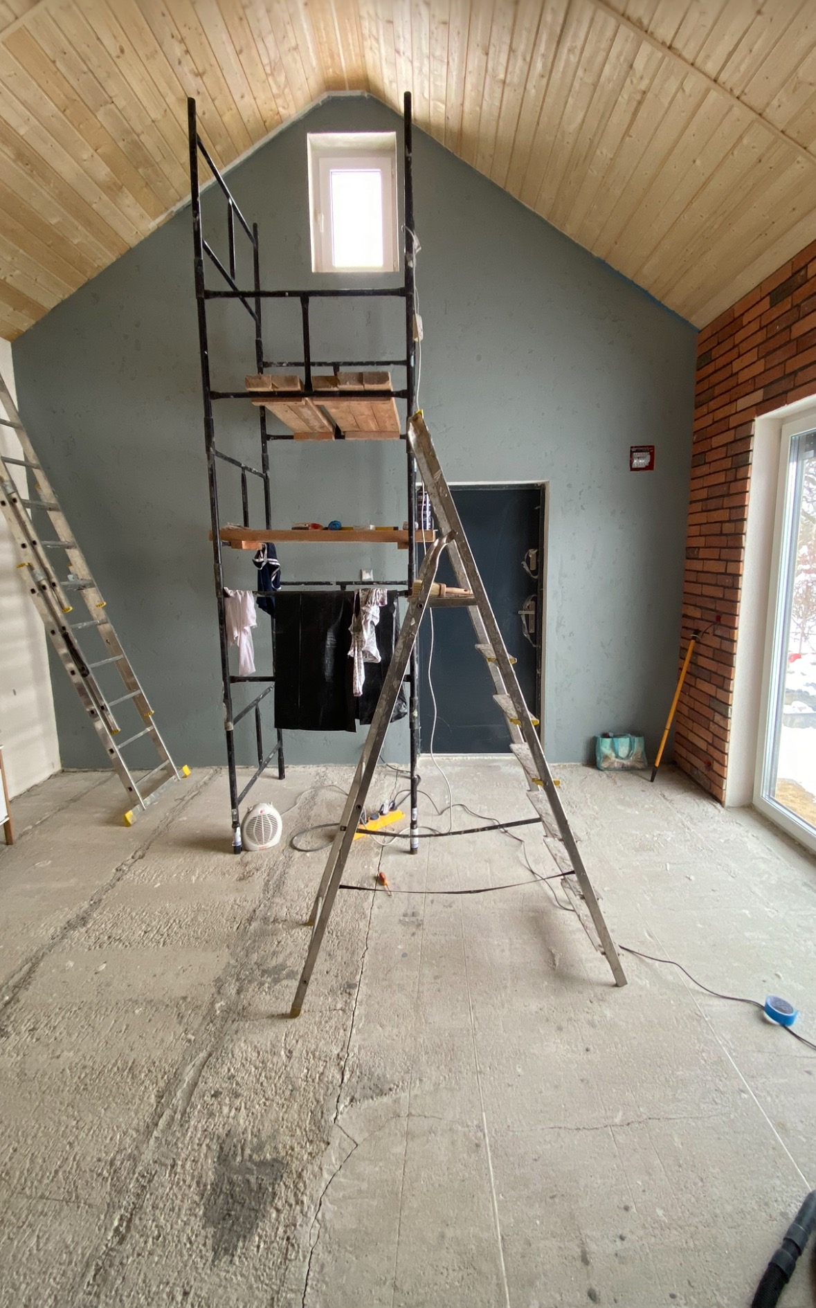 Wnętrze w trakcie remontu: surowy beton na podłodze, rusztowanie, drabina, ściana w kolorze szaro-zielonym, fragment ściany z cegły, drewniana boazeria na suficie.