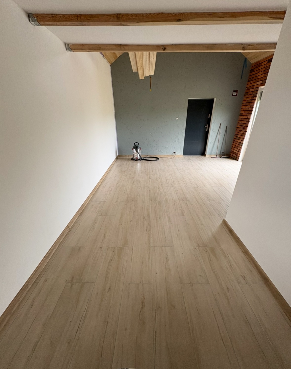 Jasne panele podłogowe w nowym wnętrzu z drewnianymi belkami stropowymi i szarą ścianą. Widoczny odkurzacz i fragment ściany z cegły.