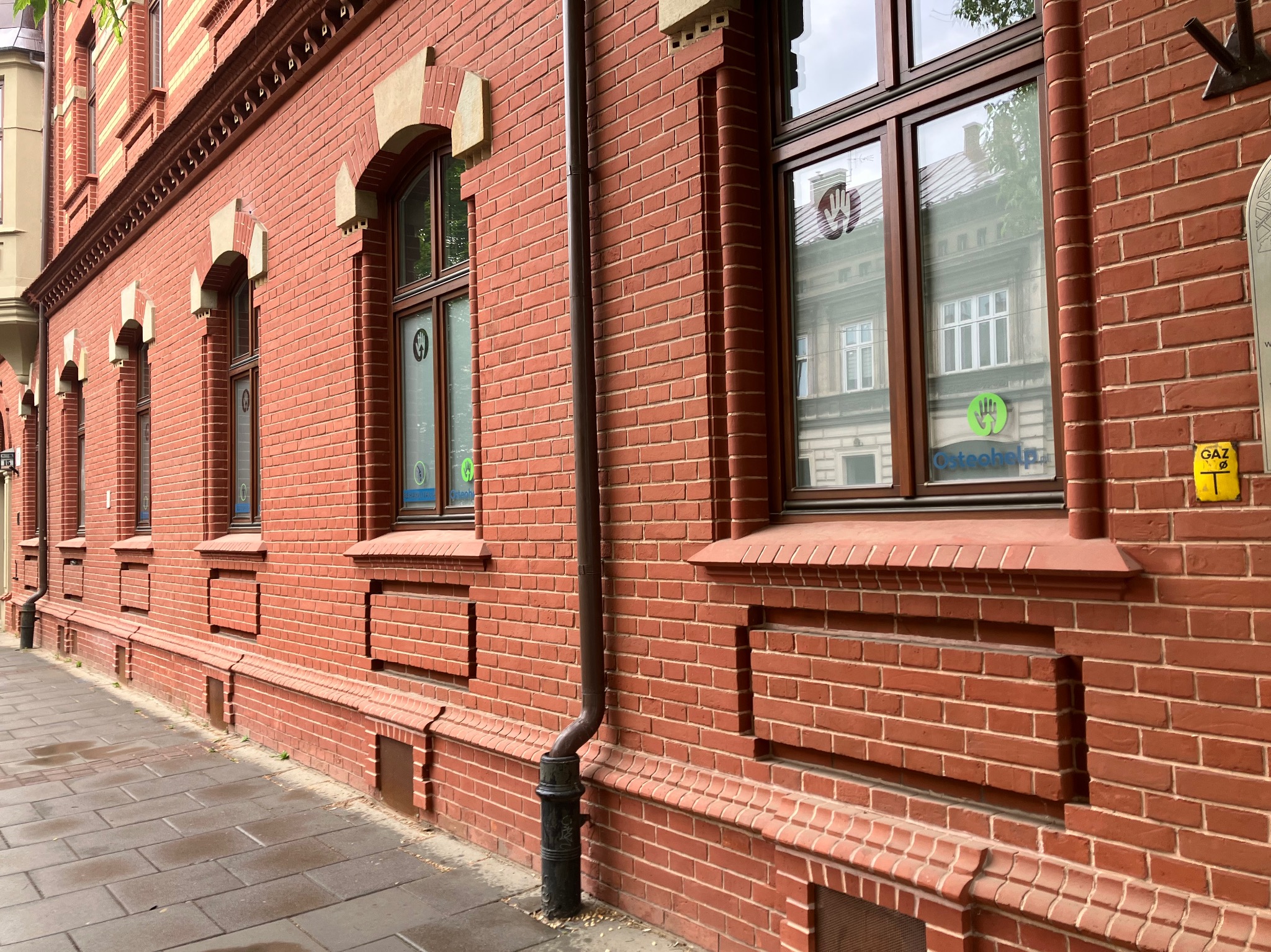 Ceglana elewacja budynku z oknami, na których widnieją naklejki z logo i adresem Osteohelp.pl ul. Limanowskiego 15, Kraków, obok żółta tabliczka z napisem GAZ.