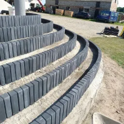 Spiralne schody ogrodowe wykończone szarymi elementami betonowymi, w tle budynki gospodarcze i prace dekarskie.