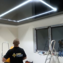 PANDA - BET DAWID ROGOZIŃSKI - Czarny sufit podwieszany z wbudowanym oświetleniem LED w kształcie kwadratu, widok z dołu, pracownik w koszulce z logo firmy montującej, drabiny i narzędzia w pomieszczeniu z oknem.
