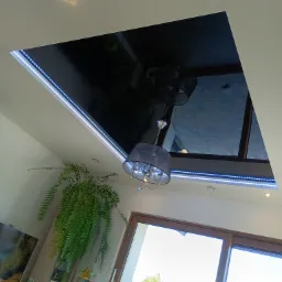 Nowoczesny salon z czarnym, błyszczącym sufitem napinanym, oświetlonym listwami LED. Widoczny żyrandol i okno z widokiem na zieleń. Nietypowe ujęcie z dołu.