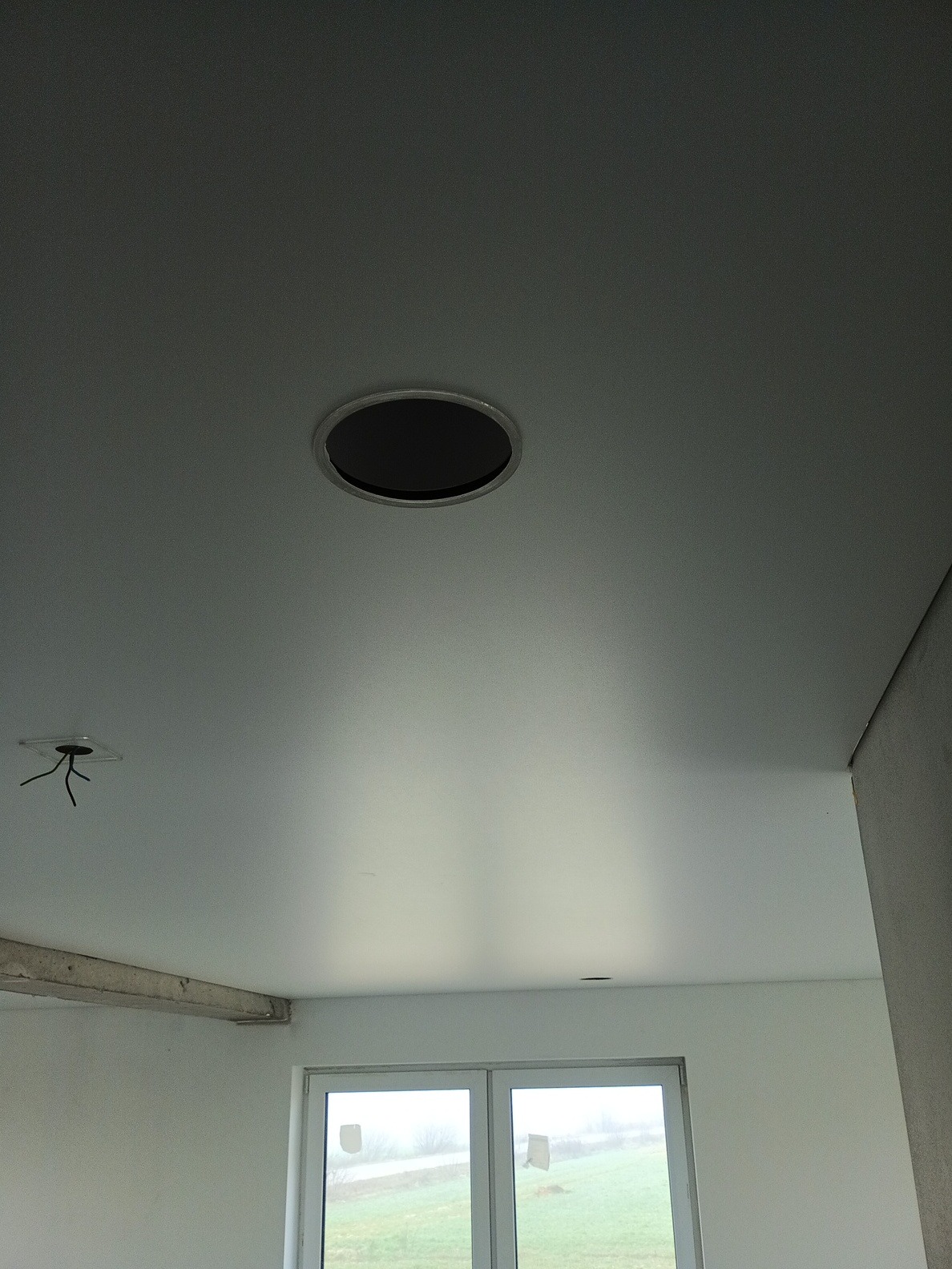 Biały sufit z okrągłym otworem na oświetlenie i wystającymi kablami, widoczne okno i fragment ściany w surowym stanie.