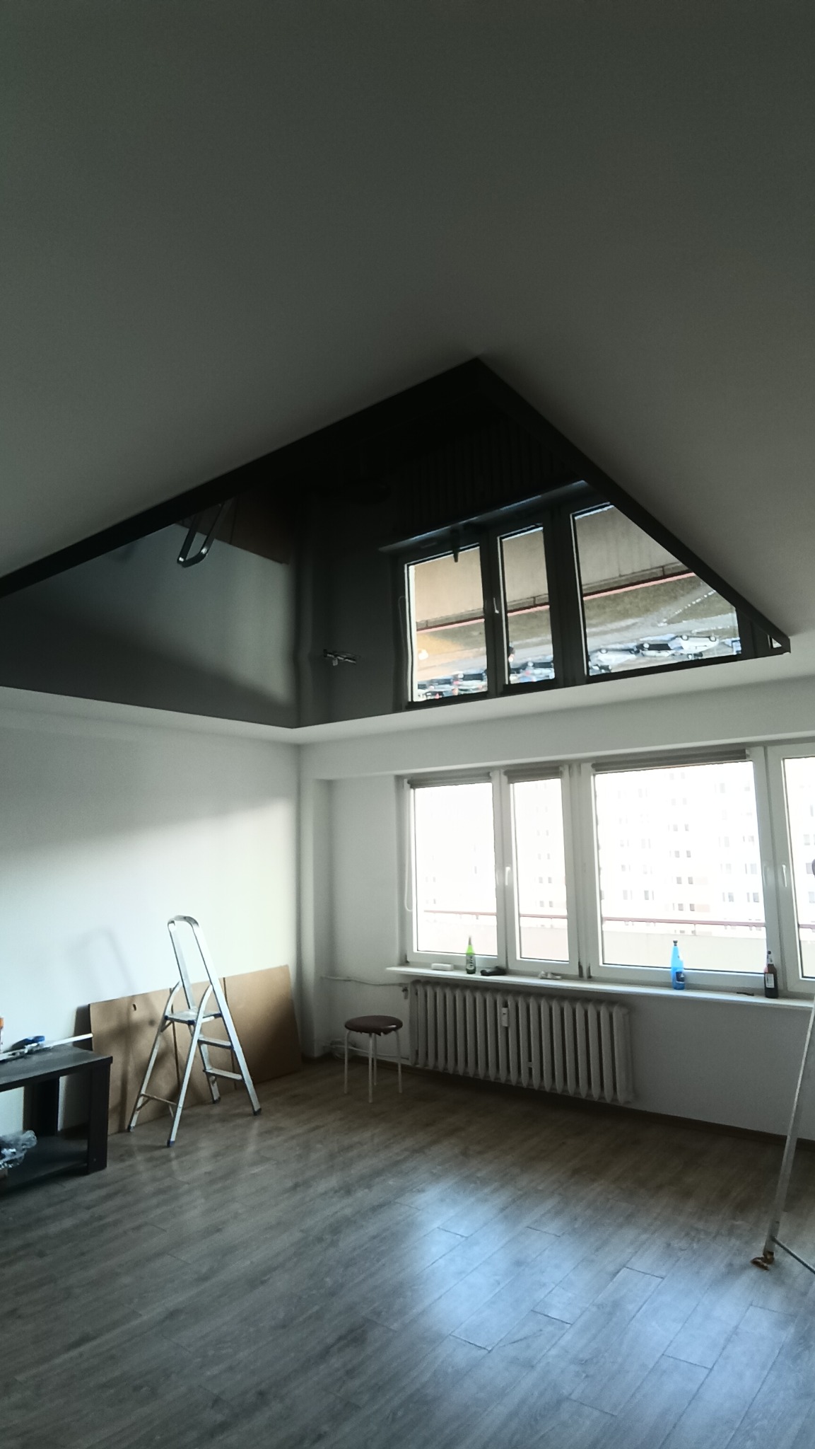Wnętrze pokoju z zamontowanym czarnym, błyszczącym sufitem podwieszanym w skośnej części dachu, drabina malarska, okno z widokiem na parking i budynek mieszkalny.