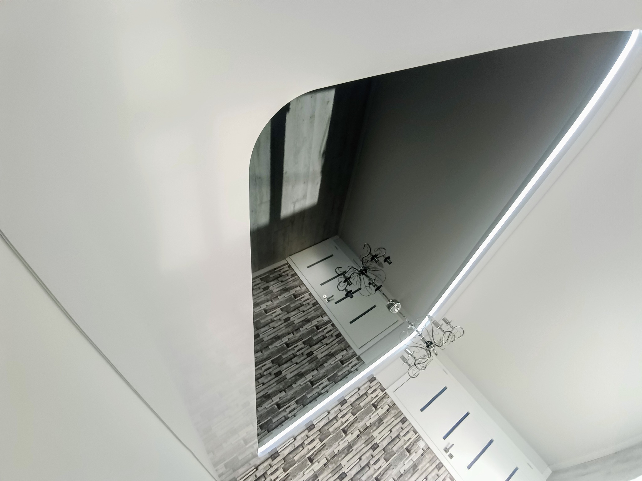 Biały, podwieszany sufit z zaokrąglonym narożnikiem i oświetleniem LED, odbijający fragment ściany z dekoracyjną okładziną imitującą kamień oraz białą drzwi.