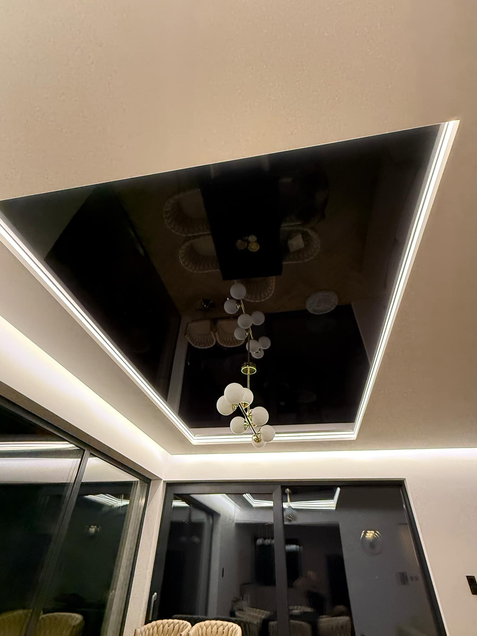 Czarny, lustrzany sufit z oświetleniem LED, odbijający wnętrze salonu z żyrandolem i widokiem na zewnątrz przez duże okna.