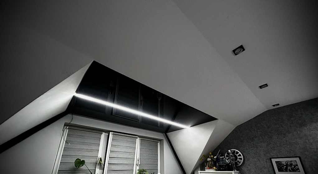 Skośny sufit poddasza z czarnym panelem i liniowym oświetleniem LED, widoczne okno z żaluzjami i dekoracje na półce.