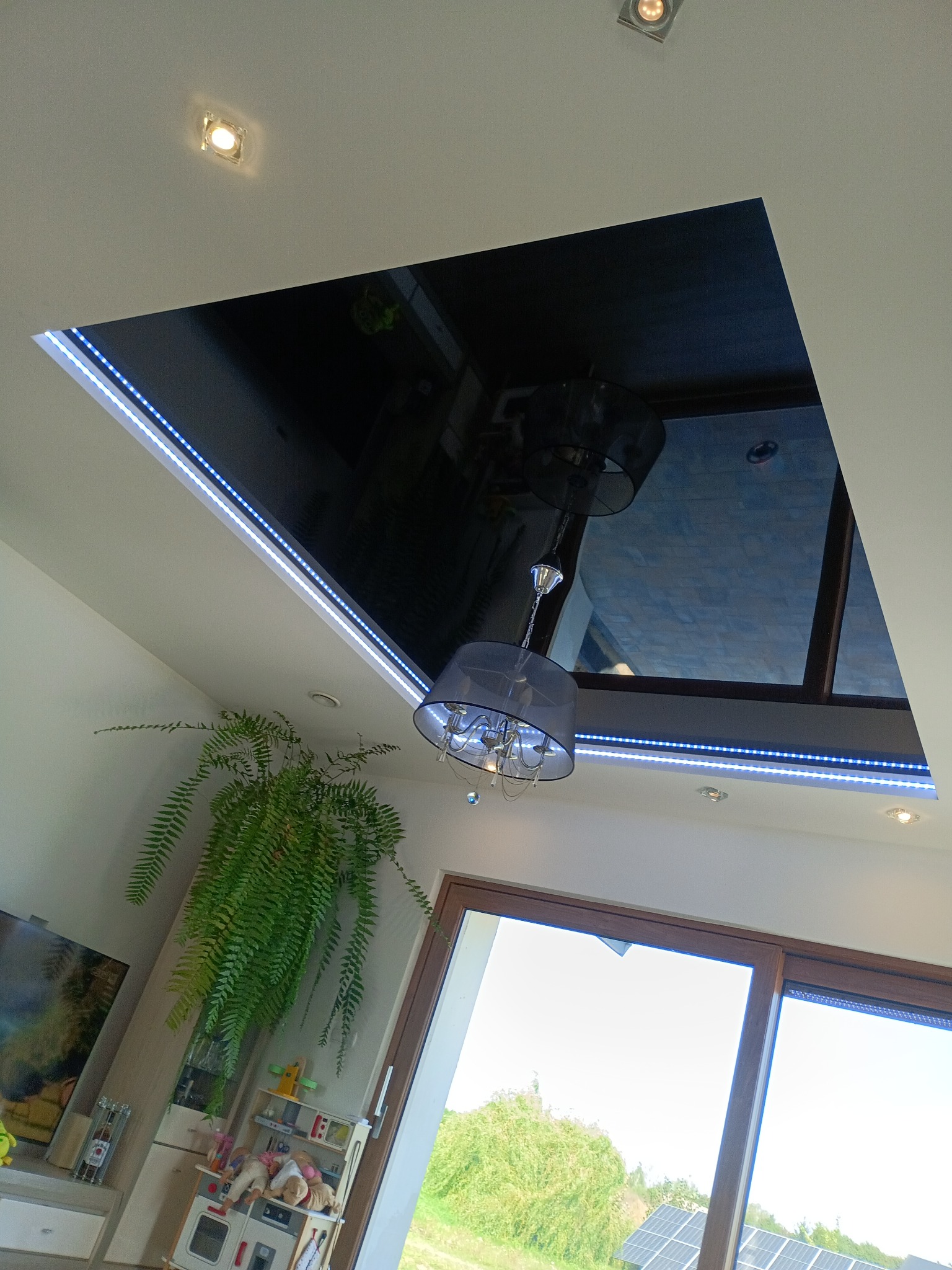 Nowoczesny salon z czarnym, błyszczącym sufitem napinanym, oświetlonym listwami LED. Widoczny żyrandol i okno z widokiem na zieleń. Nietypowe ujęcie z dołu.