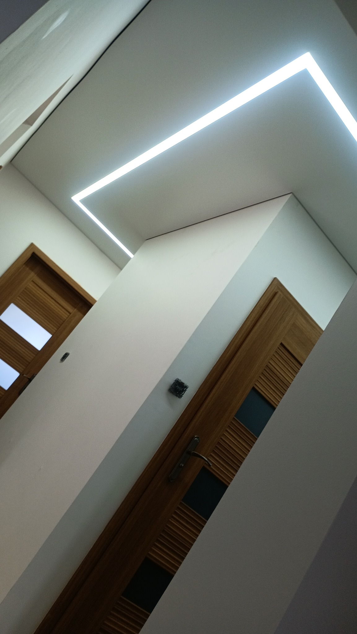Nowoczesne oświetlenie LED w kształcie prostokąta na białym suficie, widoczne drewniane drzwi z przeszkleniami. Ukośny kadr, minimalistyczny design.