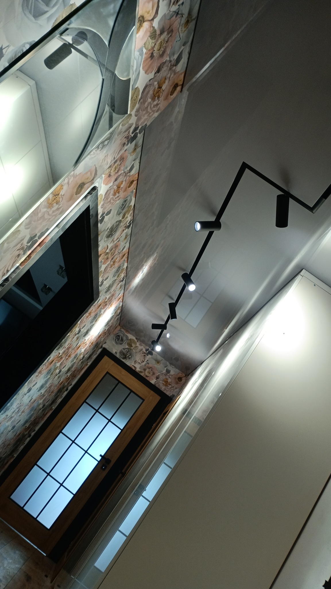 Błyszczący sufit z reflektorami, ściana w kwiaty i drewniane drzwi z szybami. Ujęcie pod kątem, ukazujące wykończenie wnętrza.