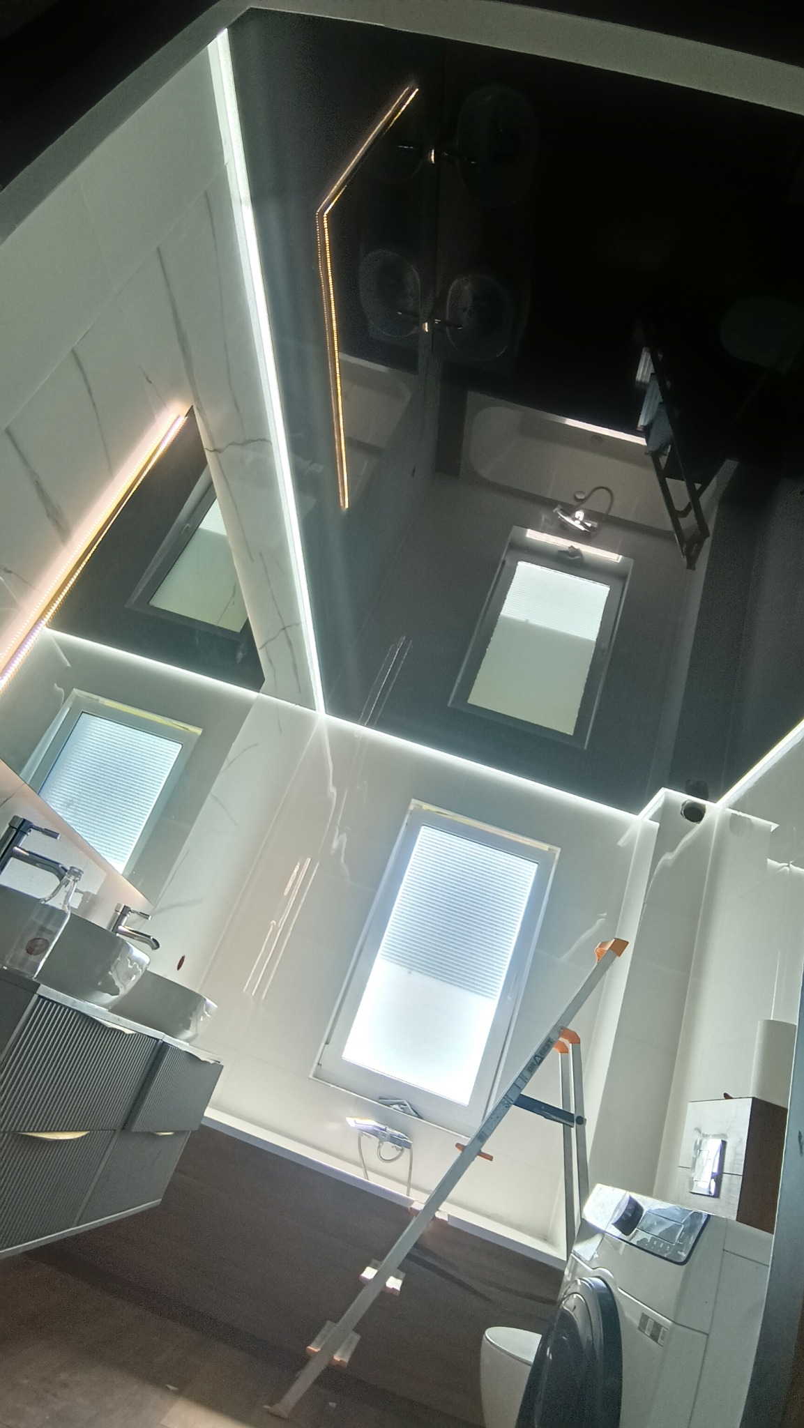 Błyszczący, czarny sufit napinany z oświetleniem LED w nowoczesnej łazience z oknami dachowymi i drabinką. Widoczna umywalka i pralka.