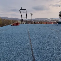 Płaski dach pokryty granulatową powłoką w kolorze niebieskim; widoczne kominy, anteny, liny i krajobraz miejski w tle.