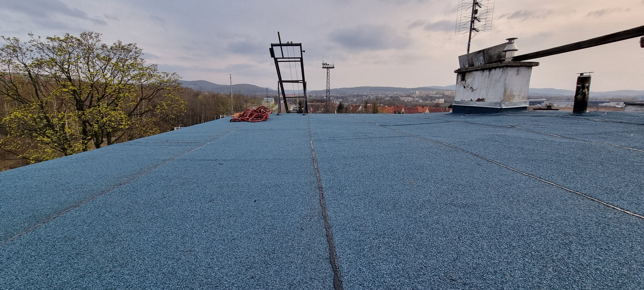 Płaski dach pokryty granulatową powłoką w kolorze niebieskim; widoczne kominy, anteny, liny i krajobraz miejski w tle.
