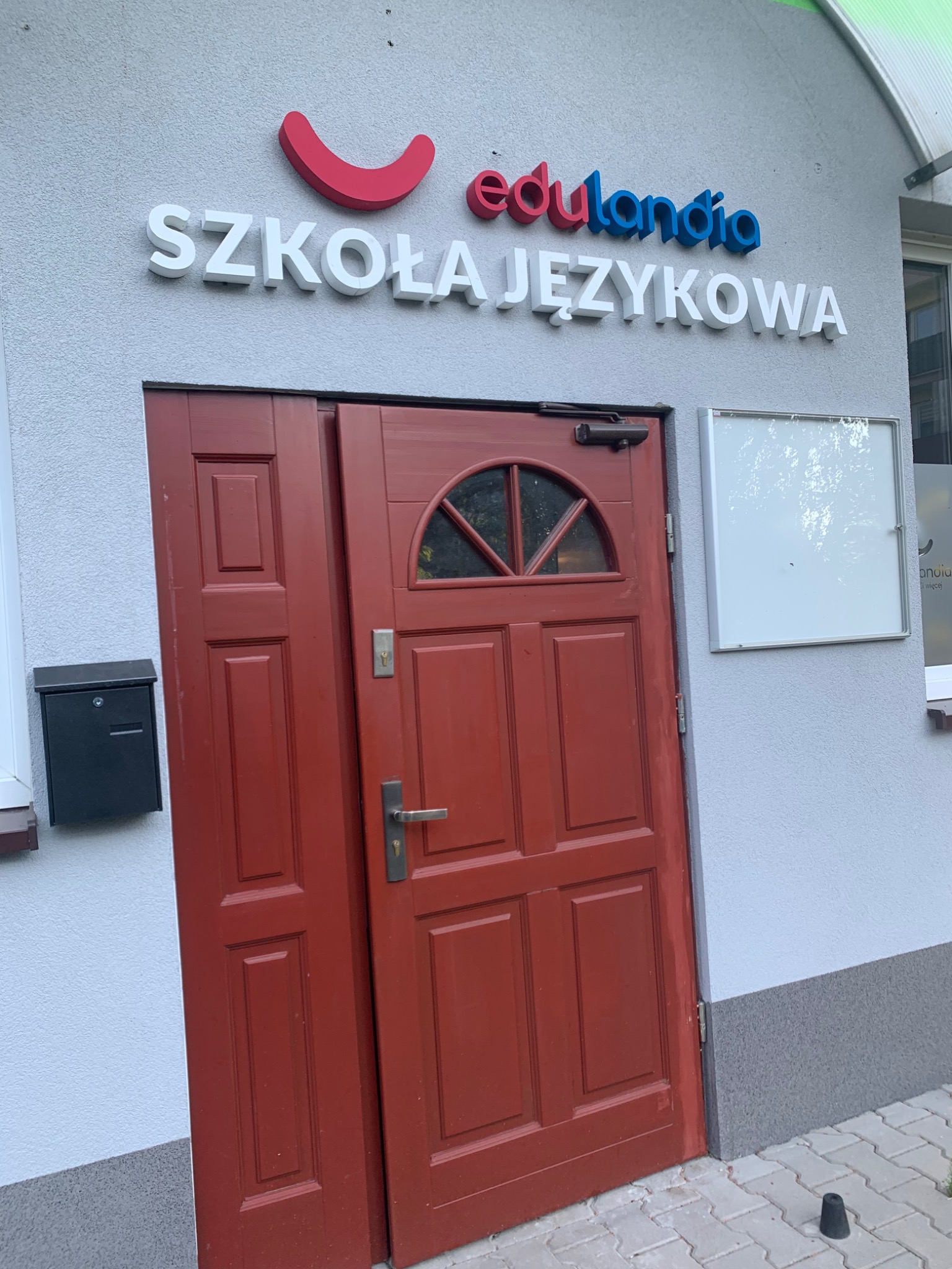 Wejście do szkoły językowej z szyldem 'edulandia Szkoła Językowa' nad czerwonymi, częściowo otwartymi drzwiami.