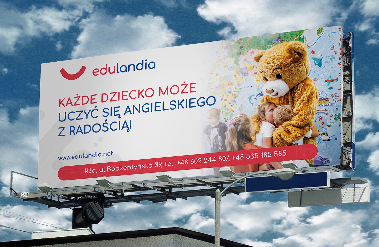Billboard reklamujący naukę języka angielskiego dla dzieci z maskotką misia przytulającego dzieci na tle mapy świata.