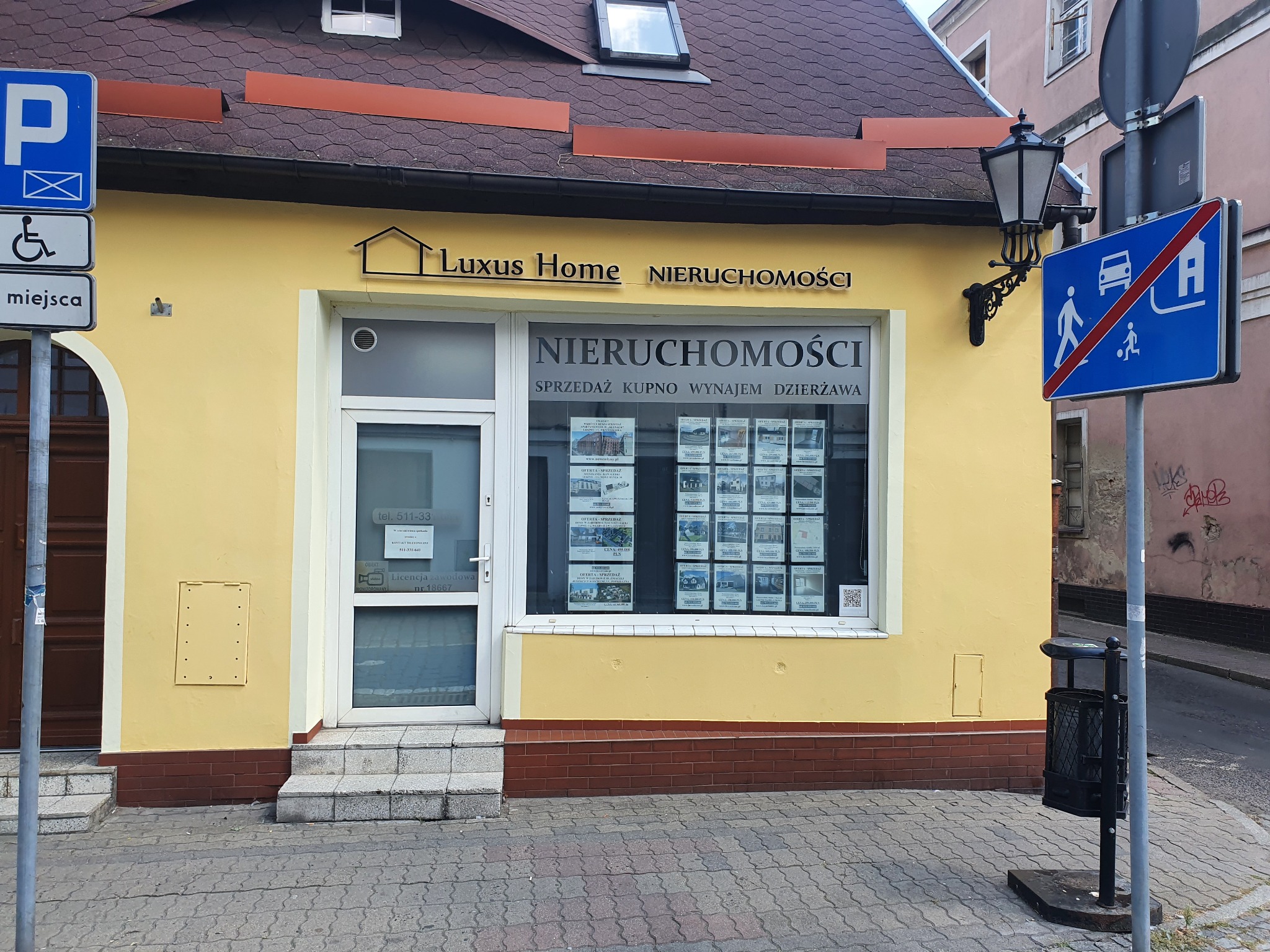 Żółta fasada biura nieruchomości Luxus Home z witryną prezentującą oferty sprzedaży, kupna, wynajmu i dzierżawy, widoczne znaki drogowe i latarnia.