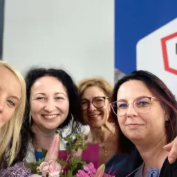 Grupowe selfie czterech uśmiechniętych kobiet z kwiatami, z logo biura rachunkowego w tle, wykonane wewnątrz pomieszczenia.