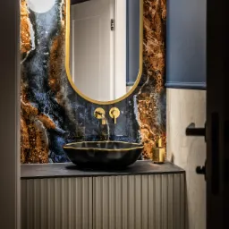 Elegancka łazienka z umywalką nablatową, złotą armaturą i lustrem w złotej ramie na tle ściany z płytek imitujących marmur.