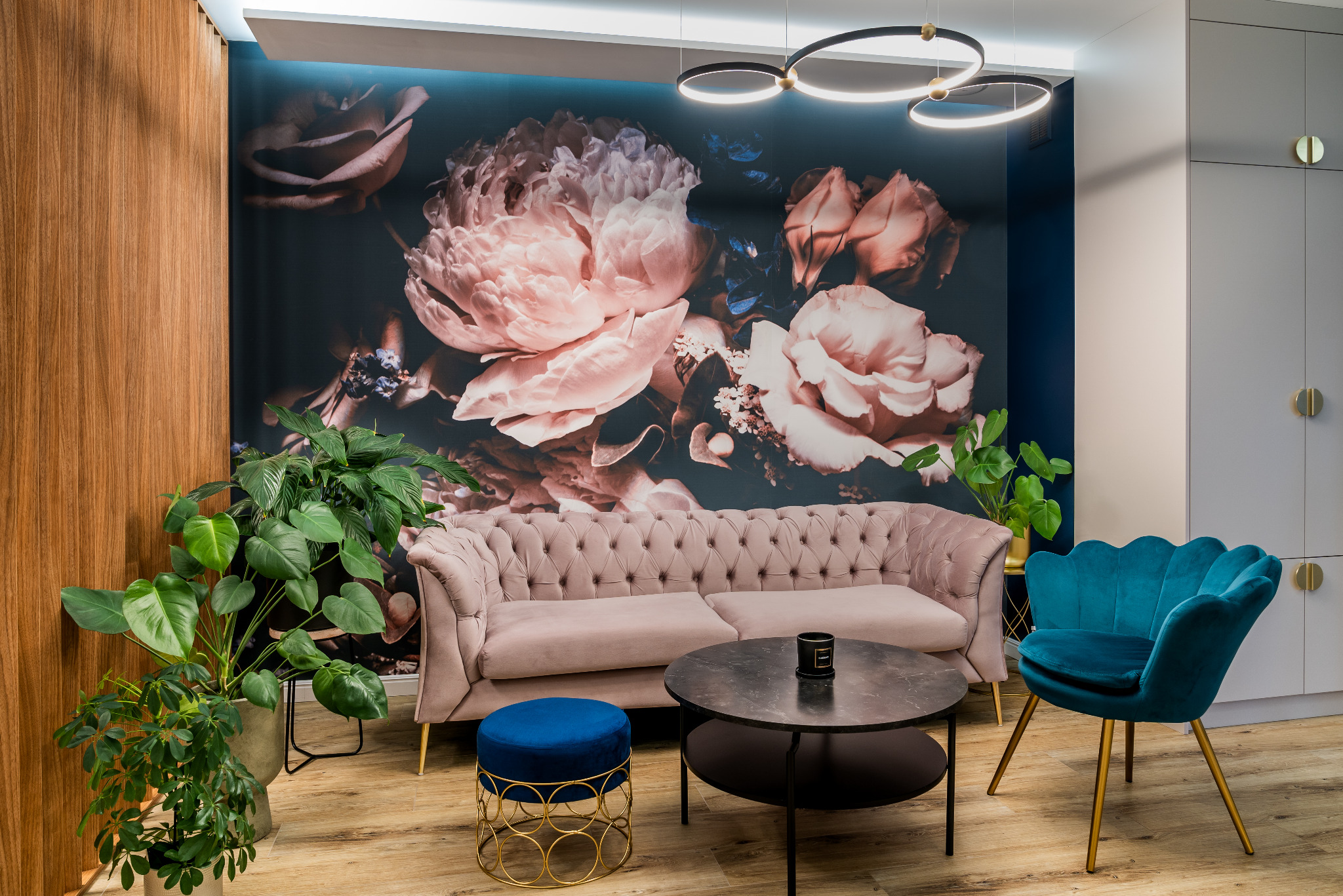 Elegancki salon z pudrowo-różową sofą Chesterfield na złotych nóżkach, okrągłym stolikiem kawowym z czarnym blatem, turkusowym krzesłem muszelką i pufą, na tle ściany z fototapetą w kwiaty.