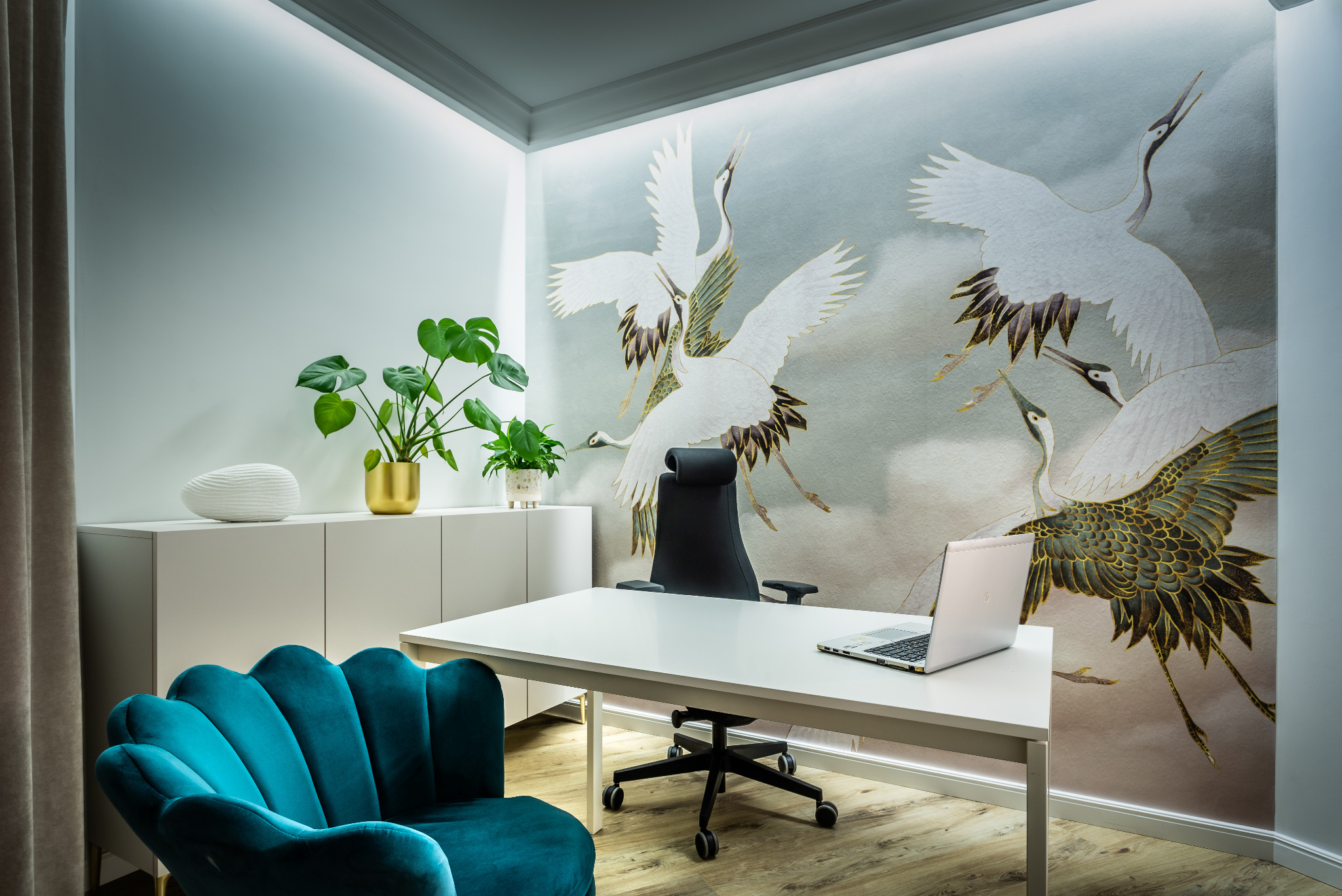 Nowoczesne biuro domowe z białą szafką, biurkiem i turkusowym fotelem na tle ściany z motywem żurawi, oświetlone listwami LED.