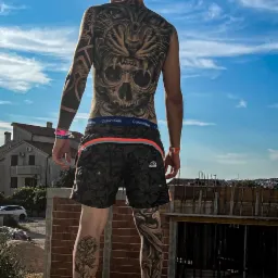 Mężczyzna z tatuażami na plecach i nogach stoi na murku z cegieł, w tle widoczne budynki i niebo.