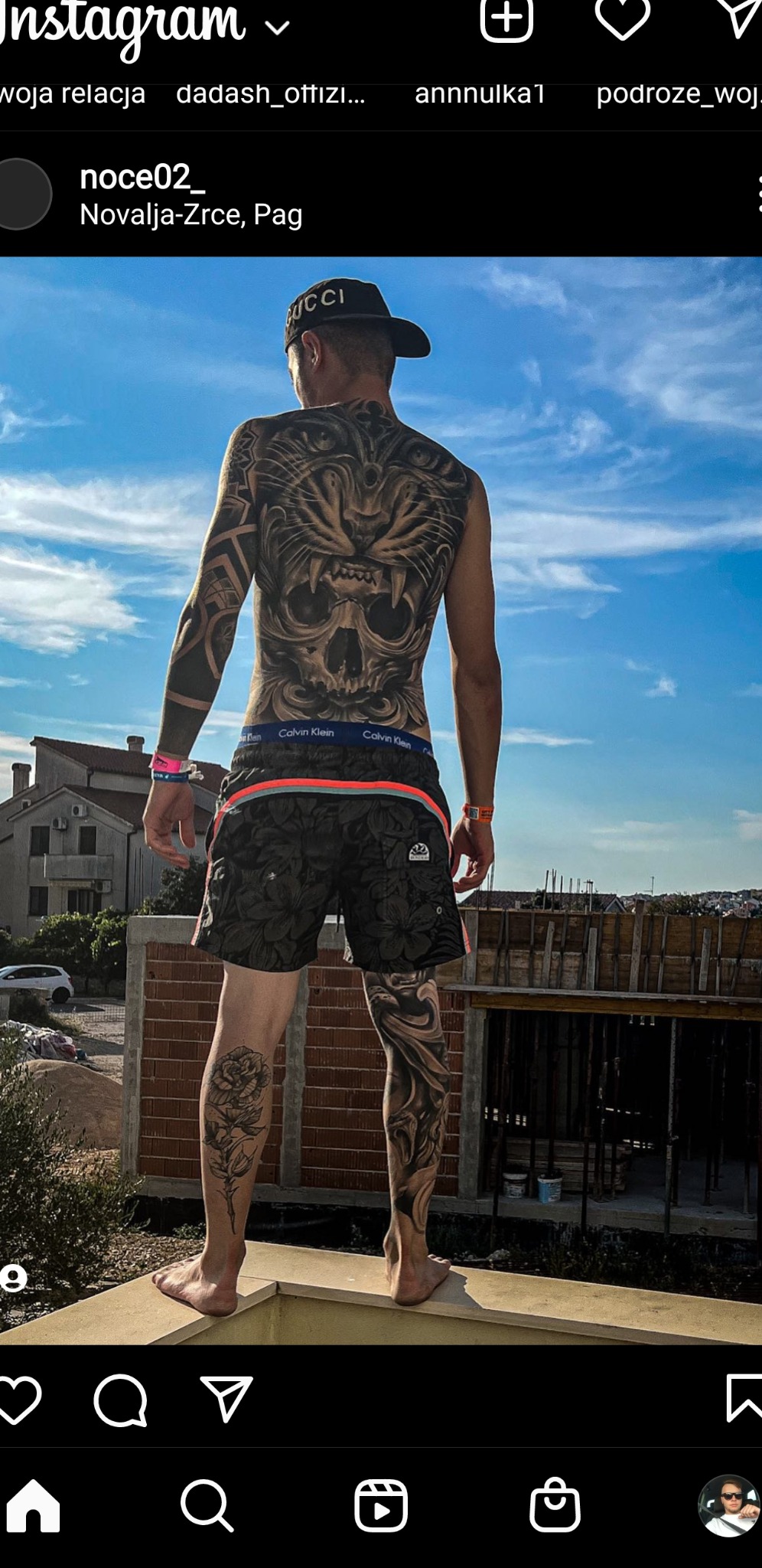 Mężczyzna z tatuażami na plecach i nogach stoi na murku z cegieł, w tle widoczne budynki i niebo.