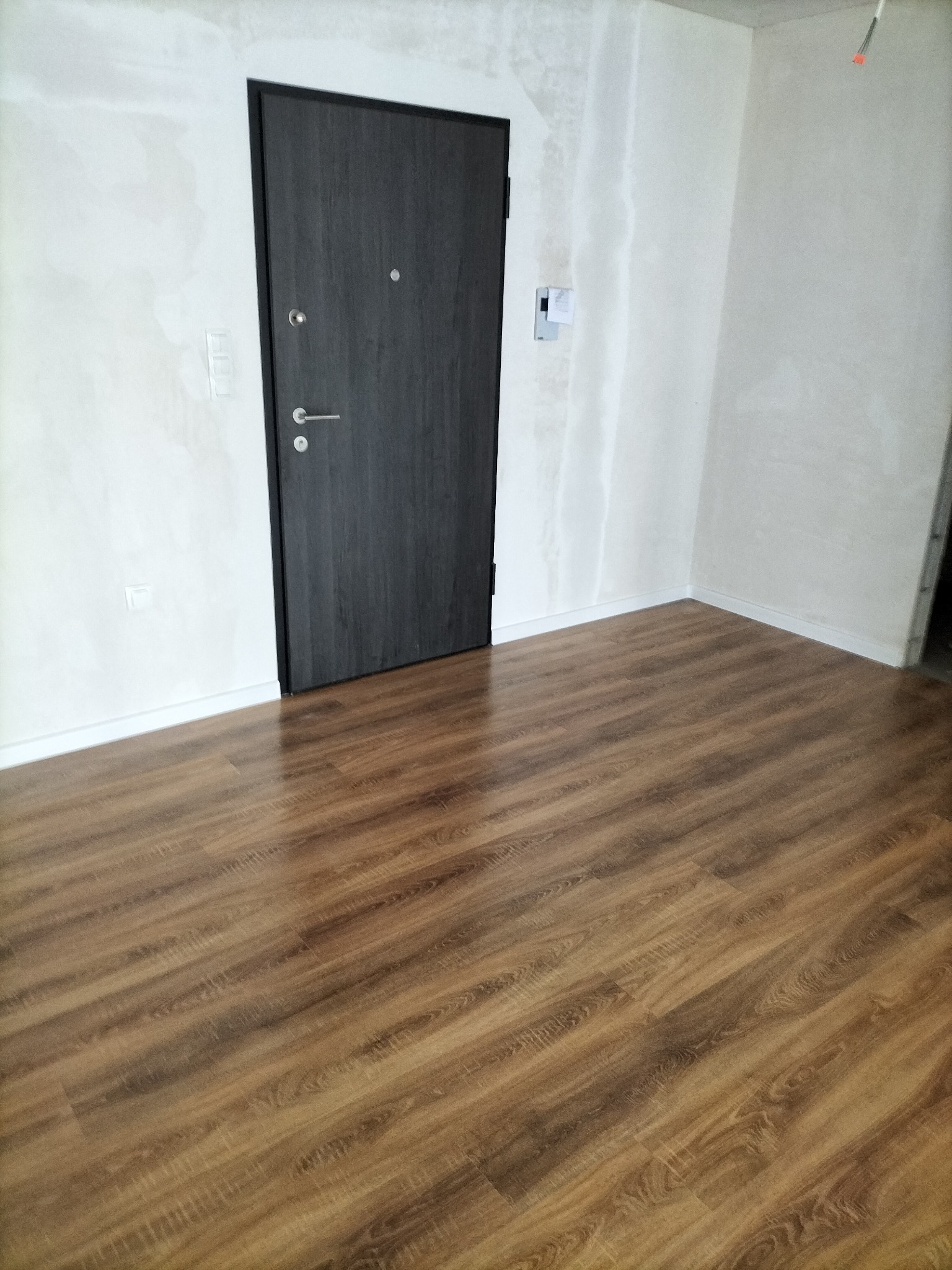 Wnętrze pokoju po położeniu paneli podłogowych w odcieniach brązu, widoczne ślady szpachlowania na ścianach i ciemne drzwi wejściowe.