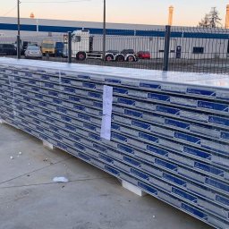 Marpanel sp z.o.o. - Paleta z dużą ilością płyt warstwowych firmy GOR-STAL, zabezpieczonych folią, na tle przemysłowego krajobrazu z budynkami i kominami.