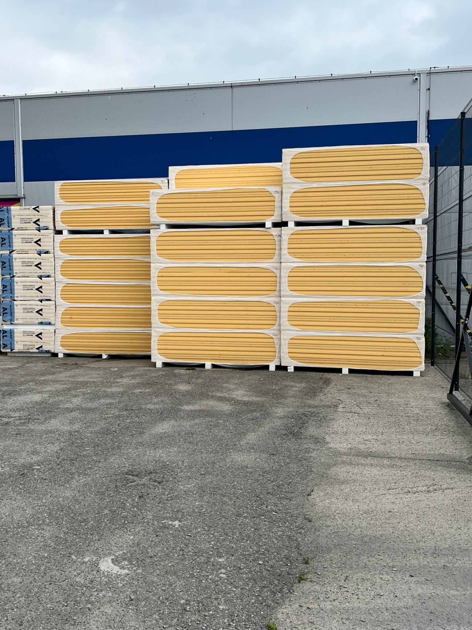 Stos żółtych płyt warstwowych TermPIR na paletach, zabezpieczonych folią, składowanych na zewnątrz budynku magazynowego z niebieskim pasem.