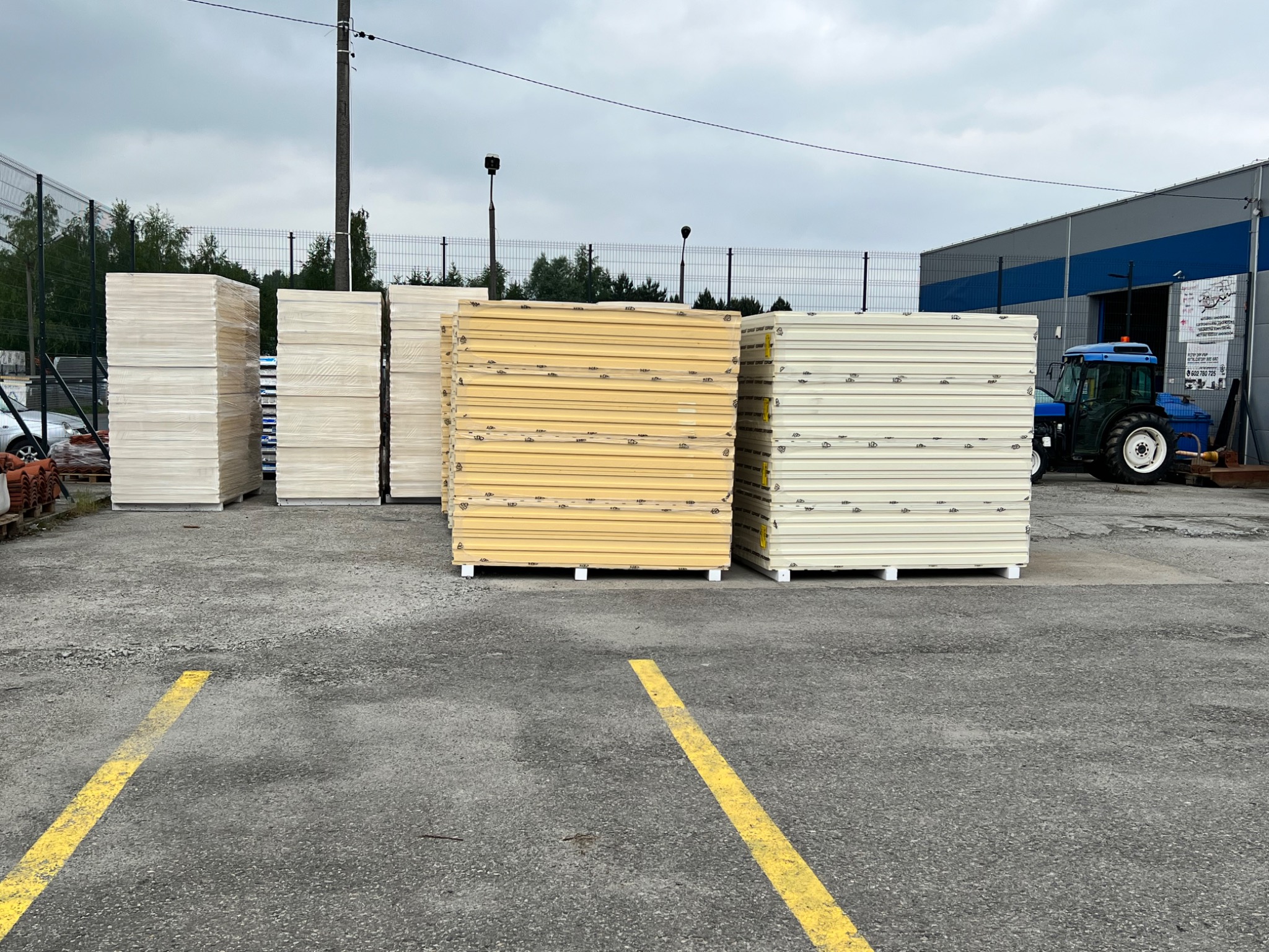 Sztaple płyt warstwowych w kolorze białym i żółtym, spakowane w folii, składowane na placu budowy, w tle widoczny budynek magazynowy i niebieski traktor.