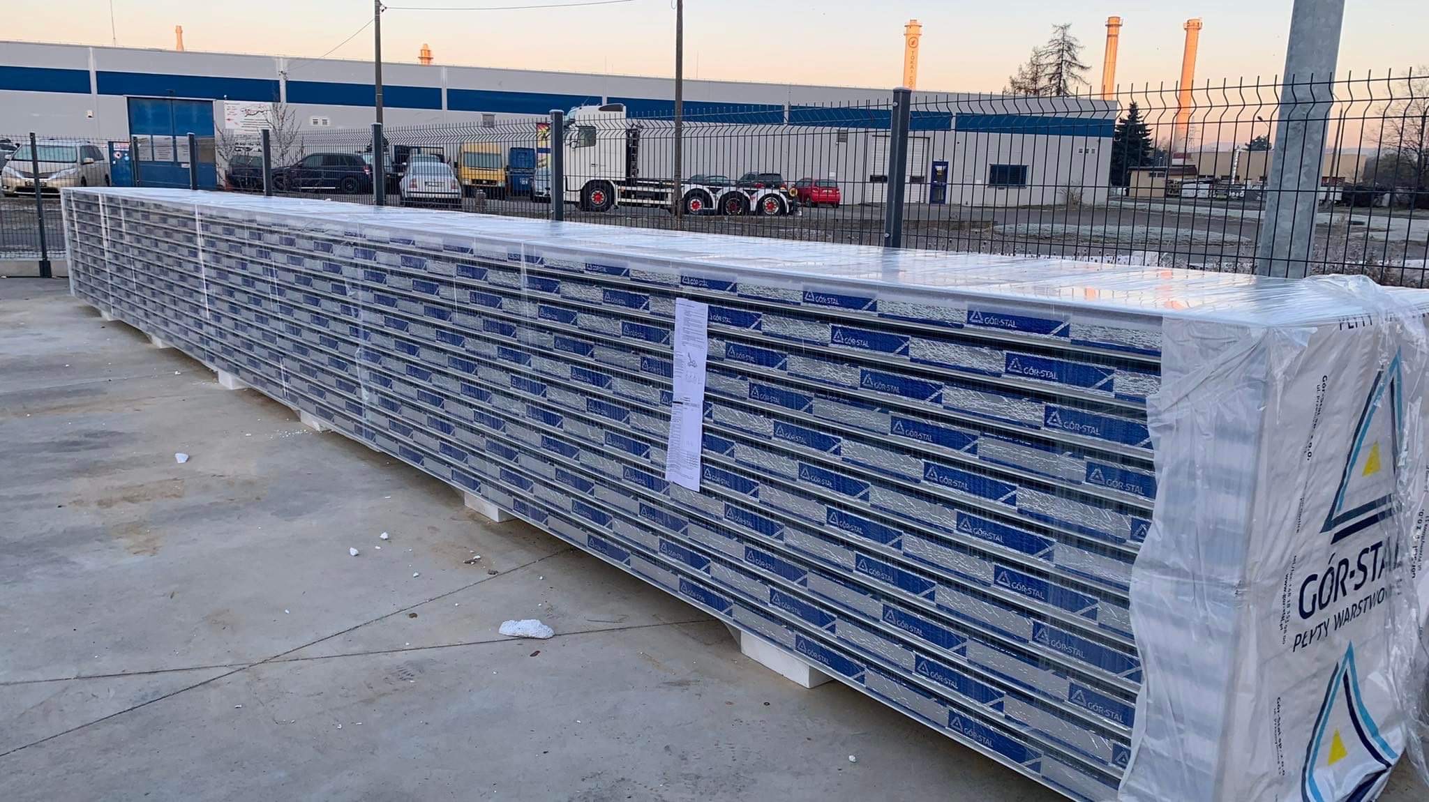 Paleta z dużą ilością płyt warstwowych firmy GOR-STAL, zabezpieczonych folią, na tle przemysłowego krajobrazu z budynkami i kominami.