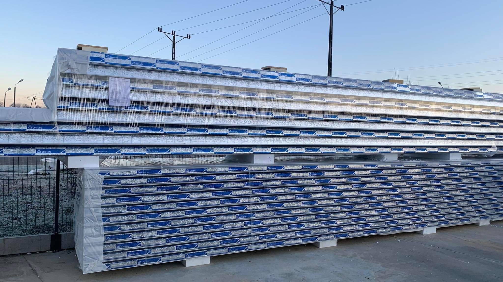 Stos płyt warstwowych z widocznym logo producenta, zabezpieczonych folią, ułożonych na paletach na zewnątrz budynku w słoneczny dzień.