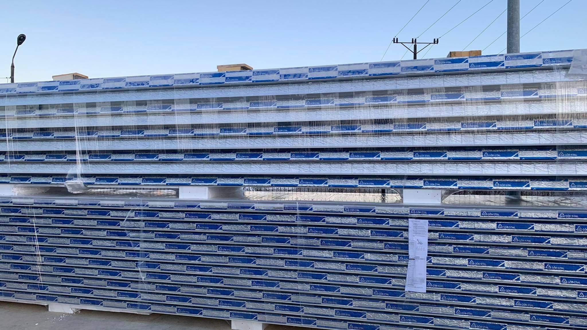 Stos płyt warstwowych z widocznym logo producenta, zabezpieczonych folią transportową, składowanych na paletach, na zewnątrz budynku.